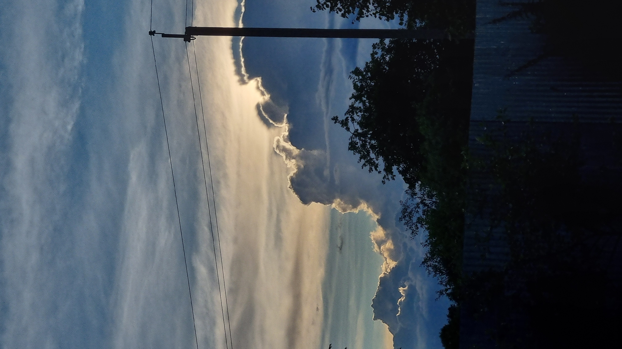 The image shows a dramatic sky with billowing clouds, possibly during sunset or sunrise. The sun is partially obscured by the clouds, creating a glowing effect along their edges, with a mix of light blue, white, and darker grey hues in the sky. A telephone pole with wires runs vertically through the left side of the frame, suggesting an outdoor, possibly rural or semi-urban, setting. To the right, silhouetted trees and foliage are visible against the sky. The far right of the image is dominated by a dark, textured surface, which appears to be corrugated metal, perhaps part of a building or fence. The overall mood is one of natural beauty and atmospheric spectacle, with the play of light and shadow adding depth and drama. There is no visible text in the image.