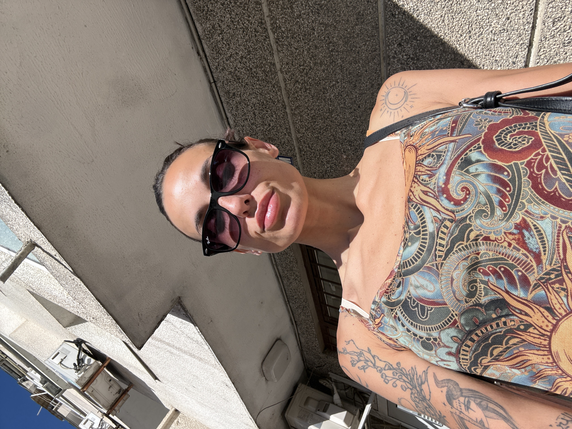 The image shows a woman standing outside a building in the city of Pleven, Bulgaria. She is wearing sunglasses and a dress with a colorful pattern. Her left arm is visible and shows several tattoos, including a bird and a crescent moon. The building behind her is made of concrete and has air conditioning units attached to it. The lighting suggests it is daytime, with strong sunlight casting shadows on the wall.