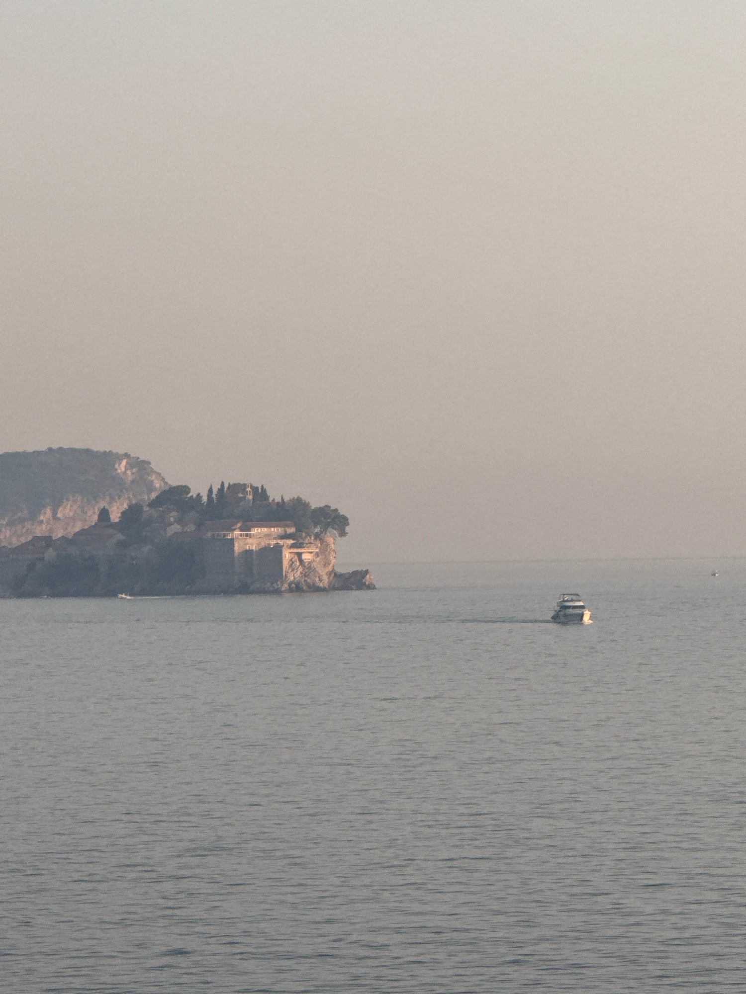 The image depicts a serene coastal scene with a prominent rocky island topped with buildings and vegetation, situated in the waters off the coast of Kamenovo, Montenegro. The island appears to be fortified, with stone structures and what looks like red-tiled roofs visible amidst the greenery. A small white motorboat is seen navigating the calm, gray water in the foreground, leaving a subtle wake. The sky is hazy and a pale, muted color, suggesting either early morning or late afternoon light. The overall atmosphere is tranquil and somewhat muted due to the atmospheric conditions.