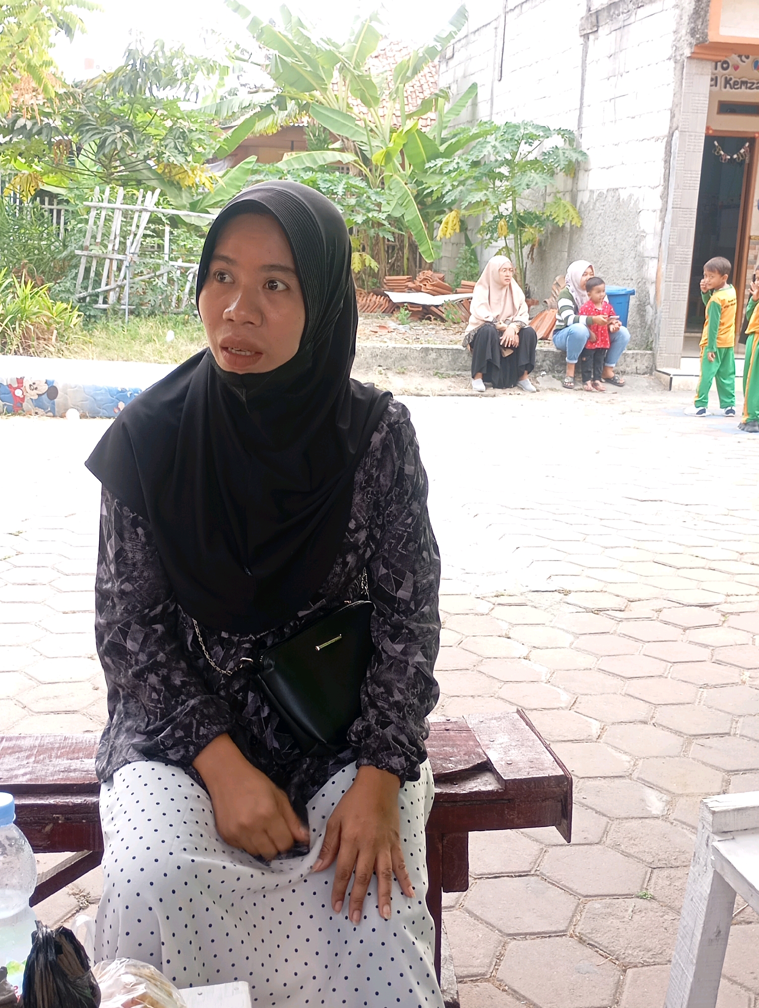 In Babelan, Indonesia, a woman wearing a black hijab and a patterned black and white dress sits on a wooden bench, facing left. Her left hand rests on her lap, while her right hand is near her waist. A black purse with a silver chain strap is visible on her left hip. To her right, a bottle of water and a black plastic bag are on the bench. In the background, a group of people, including children in school uniforms, are gathered near a building. The ground is paved with hexagonal tiles. Lush green foliage, including banana trees, is visible on the left side of the image.