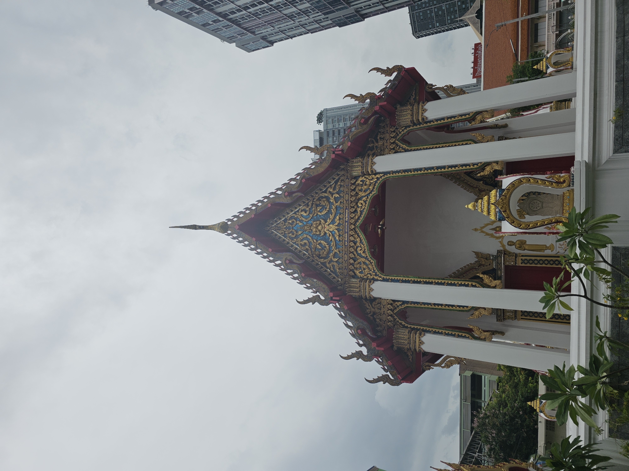 This is a low-angle, full shot of a Buddhist temple pavilion in Bangkok, Thailand. The sky is overcast and grey. The pavilion is white with ornate gold and blue trim along the roof. The roof is tiered and has decorative finials. There are large white pillars supporting the roof. Behind the pavilion, there are modern high-rise buildings visible. In the foreground, there are green plants. The pavilion appears to be empty.