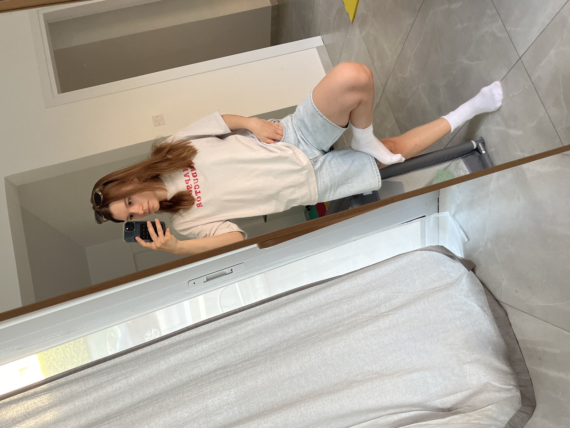 A young woman with auburn hair is taking a mirror selfie. She is wearing a light-colored t-shirt with red lettering that appears to be reversed, denim shorts, and white socks. She has sunglasses pushed up on her head and is resting her right leg on a stroller. The scene is indoors, with a large mirror reflecting the woman and a doorway in the background. A light-colored curtain hangs to the left, partially obscuring a window with sunlight streaming through. The floor is tiled in a light grey color. This scene is in Limassol, Cyprus.