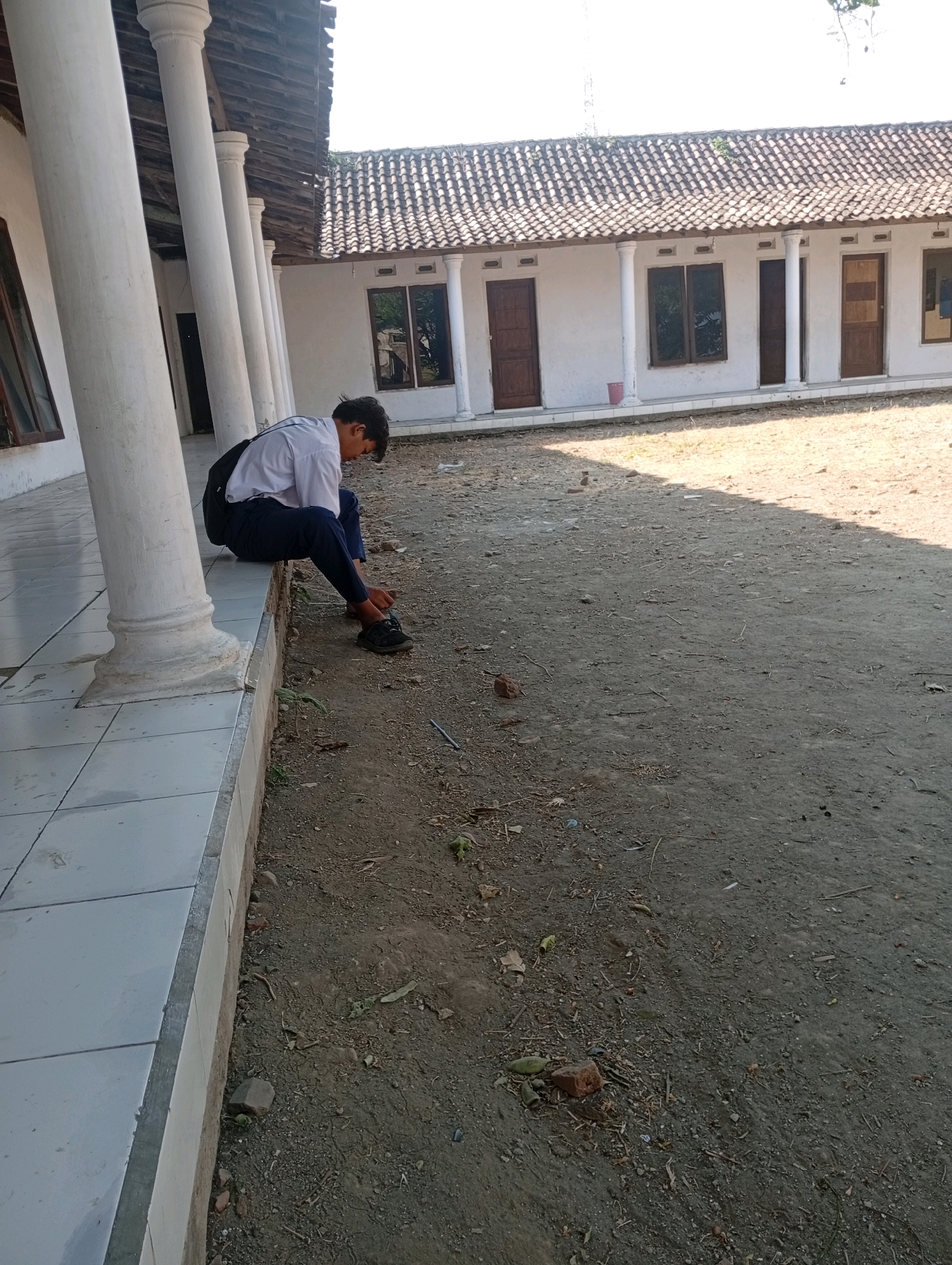 A lone young male, wearing a white school shirt and dark pants, is seated on a raised tiled platform next to large white pillars. He is facing down, appearing to tie his dark sneakers. A black backpack is slung over his left shoulder. The setting is outdoors, with a dirt and gravel ground. Behind him is a building with a tiled roof, white walls, and several windows and doors. This location is identified as Gempeng, Indonesia.