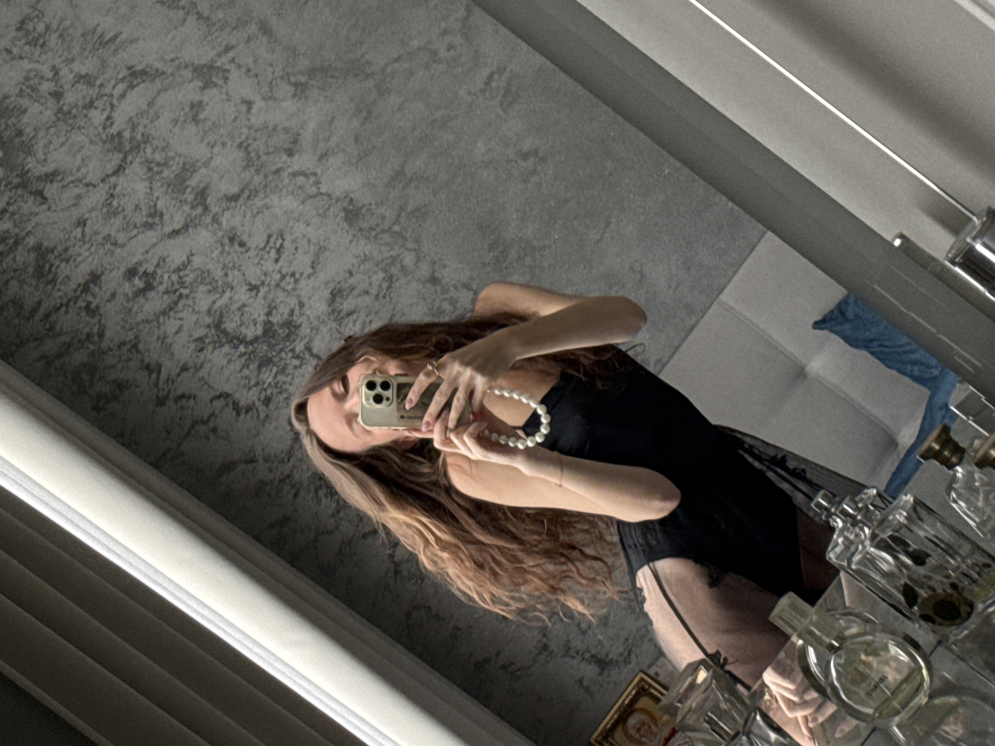 A mirrored selfie of a woman, head and shoulders, in what appears to be a bathroom setting. The woman is holding a smartphone with both hands, partially obscuring her face, while wearing a black, low-cut garment. A string of white pearls is draped around her fingers. Her long, wavy hair falls down her shoulders. The background includes a textured gray wall on one side and a partially visible interior with a blue towel or garment, suggesting a bathroom or dressing area. A collection of perfume bottles and a small framed picture are visible on a reflective surface, presumably a shelf or vanity. The photo is taken at an angle, giving a skewed perspective of the scene.