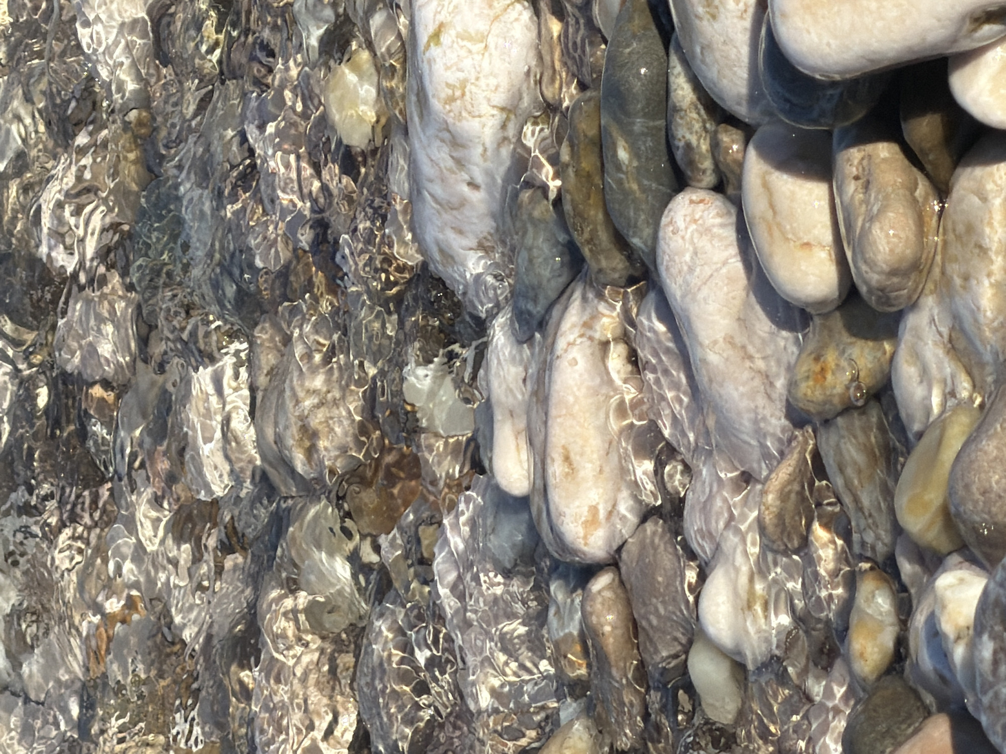 The image depicts a close-up view of a rocky shoreline, likely in Općina Baška, Croatia, with shallow, clear water flowing over the stones. The rocks are of various sizes, shapes, and colors, ranging from smooth, rounded pebbles to larger, irregular stones. They are a mix of grays, browns, whites, and earthy tones. The water creates rippling reflections and refractions on the surfaces of the rocks, giving the scene a dynamic and glistening appearance. The texture of the rocks is varied, with some appearing smooth and polished by the water, while others have more rough and defined edges. The overall impression is one of a natural, serene coastal environment. There are no people or animals visible in the image. The focus is entirely on the geological and aquatic elements of the scene.
