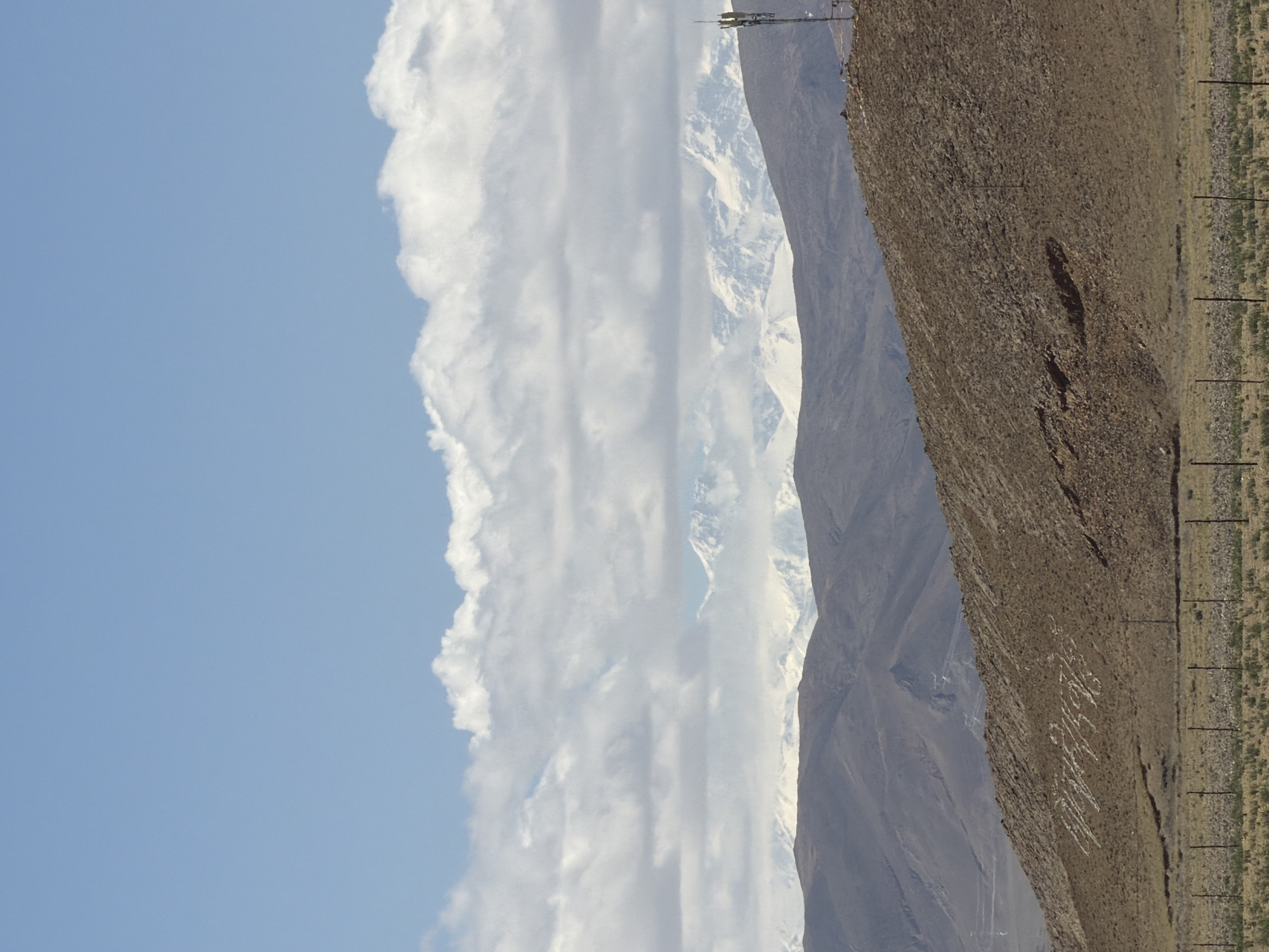 The image depicts a vast mountain landscape under a partially cloudy blue sky, captured during daytime. The original image is oriented vertically, but the content suggests a horizontal landscape view.
In the upper portion of the frame, a clear blue sky dominates the left side, meeting a long, thick band of white, cumulus clouds that stretch horizontally across the middle of the sky. Behind this cloud bank and extending into the background are majestic, snow-capped mountain peaks, indicating a high-altitude or cold region. A communications tower or mast is visible on one of the distant mountain ridges, just above the snowline.
The lower right half of the image is dominated by a barren, brown, rocky, and sandy foreground slope. This slope appears arid with minimal vegetation. Prominently written in white characters on this brown slope is Chinese text, which reads "中国最美" (Zhōngguó zuìměi), meaning "China's most beautiful". To the far right of the foreground, a strip of sparse, dry vegetation is visible, along with several thin, dark vertical posts, possibly fence posts or utility poles, extending into the distance. The overall scene suggests a rugged, remote mountain environment, consistent with the location context of Mainpu township, China.