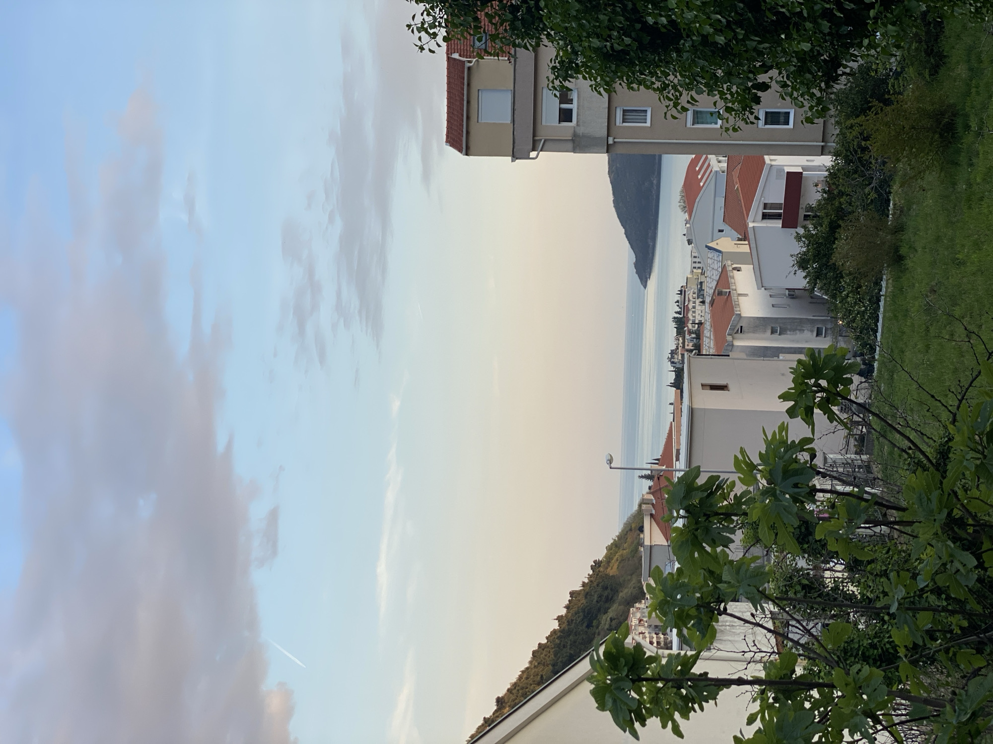 The image depicts a view of Budva, Montenegro, from an elevated position. The sky is visible with a mix of clear blue and cloud cover. In the distance, a mountain or large hill can be seen across the water. The middle ground consists of buildings of varying heights and architectural styles, many with red-tiled roofs. Green foliage and trees are present in the foreground and mid-ground, partially obscuring some of the buildings. A street lamp is visible amongst the roofs. The overall scene suggests a residential area with a coastal view.