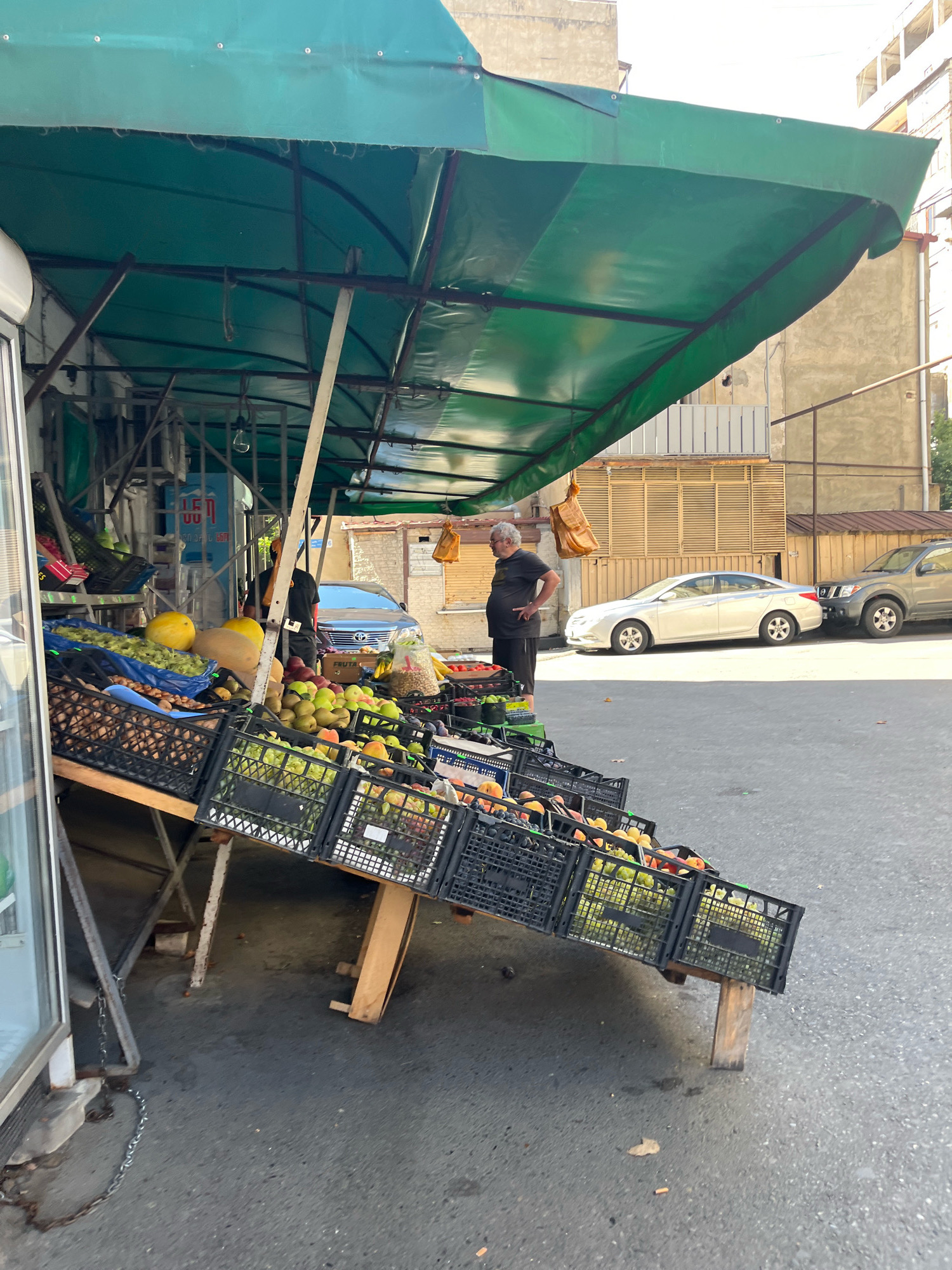A street market stall displaying fruits and vegetables is set up outdoors on a paved road. The stall has a green tarp providing shade, supported by metal poles and wooden beams. The produce is arranged in black plastic crates, which are stacked and tilted on wooden supports, creating a tiered display. Among the visible items are yellow melons, green and red fruits resembling apples or pears, and clusters of grapes. A man with graying hair and a dark t-shirt and shorts stands behind the stall, facing away from the camera. In the background, a light-colored sedan and a darker SUV are parked on the street. Behind the vehicles, buildings with textured walls and balconies are visible. The scene appears to be in an urban setting, likely in Tbilisi, Georgia, judging by the architecture and the style of the street market.
