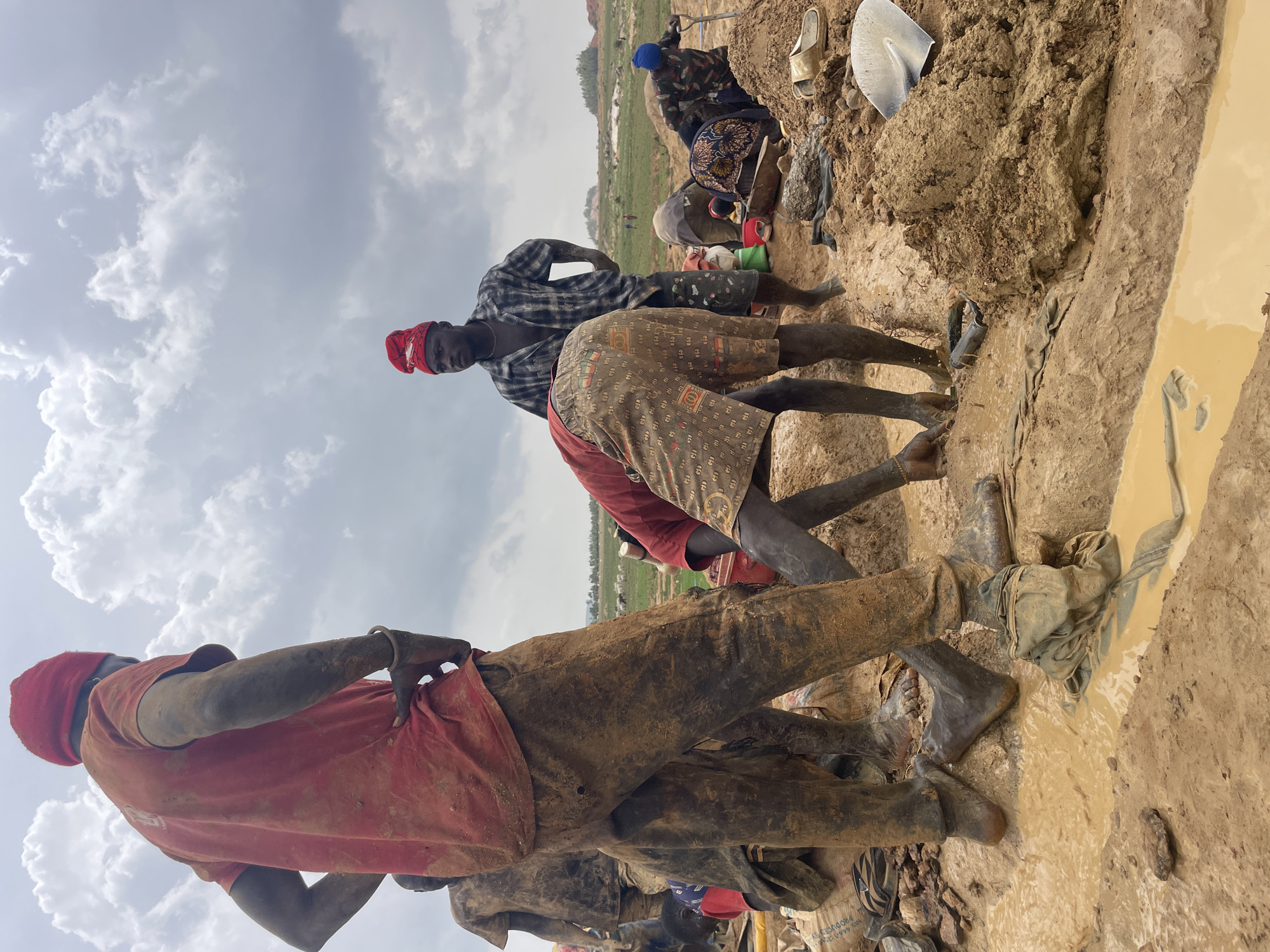 The image depicts a group of individuals engaged in labor in a muddy environment, likely a mining site, near the city of Maiyanga, Nigeria. The workers are covered in mud, suggesting difficult working conditions. At least four people are visible, wearing casual clothing and head coverings. They are positioned around a trench filled with muddy water. A shovel and other unidentified objects are scattered nearby. The background shows a partially cloudy sky.