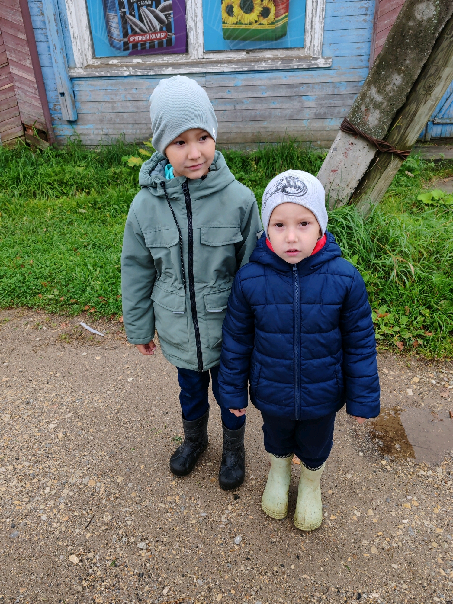 Two young children, a boy and a girl, stand on a dirt path in front of a weathered wooden building. The boy, on the left, wears a light green jacket and a grey hat. The girl, on the right, is dressed in a dark blue puffy jacket and a grey hat with a graphic design. Both children are wearing boots; the boy has black rubber boots, and the girl wears light green rubber boots. The building behind them has blue and red wooden siding. A portion of the building's window shows a poster featuring sunflowers. A leaning wooden post, bound with wire, is visible to the right of the children. Green grass grows along the side of the path and around the building. The ground appears damp in places, suggesting recent rain.