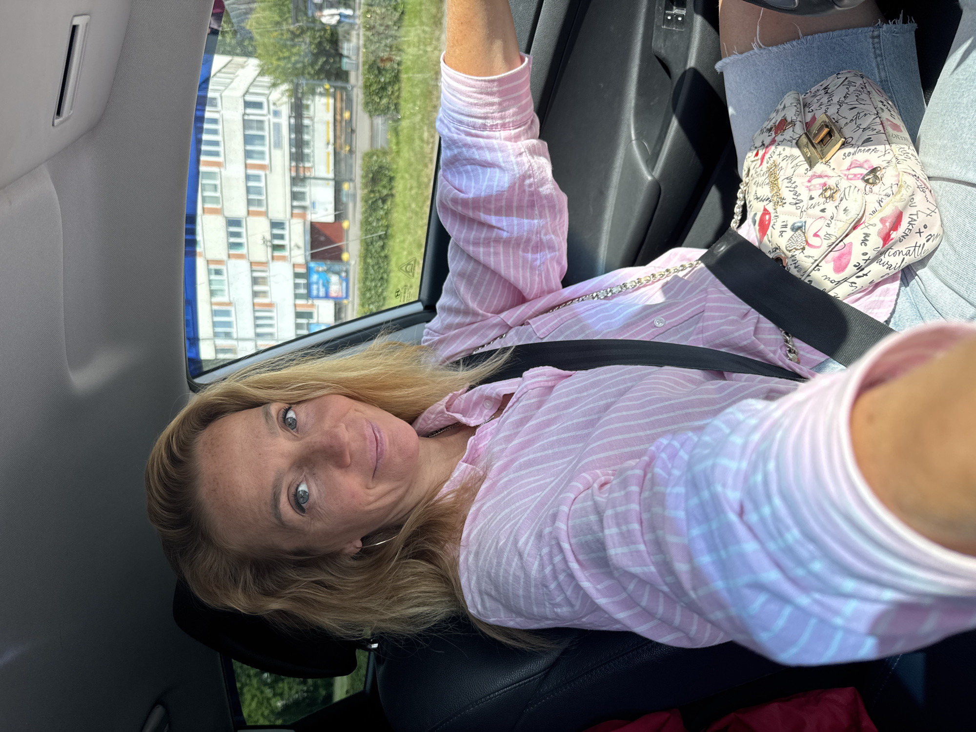 A blond woman with freckles is sitting in the driver's seat of a car, taking a selfie. She is wearing a pink and white striped button-down shirt and a denim skirt. She has a patterned purse on her lap, and a seatbelt across her chest. The car's interior is visible, including the dashboard and a portion of the passenger seat. Outside the car, a multi-story apartment building and trees can be seen, indicating an urban or suburban environment. The setting is likely Sinyavino, Russia, based on the architecture and vegetation.