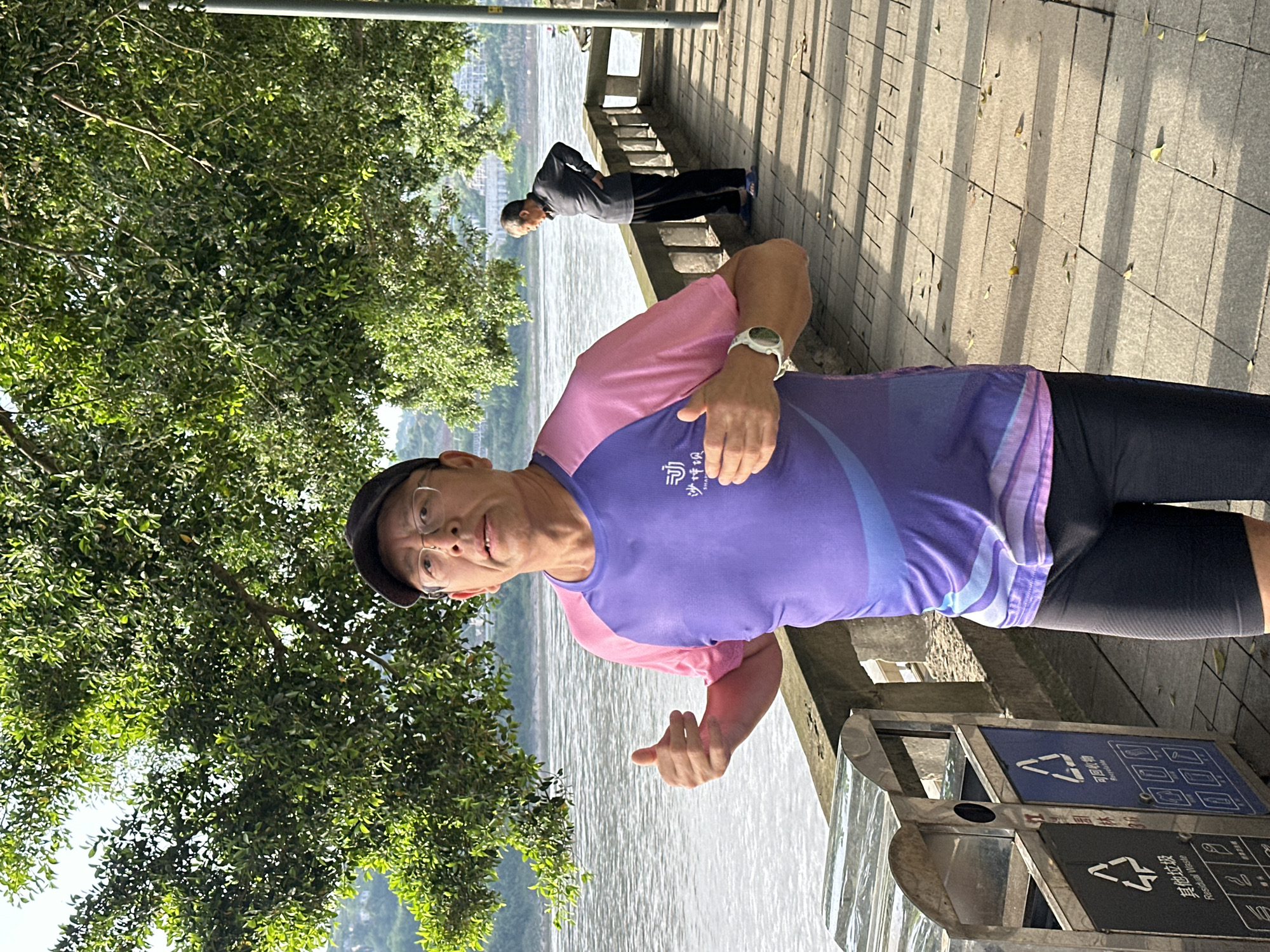 This image captures a vibrant outdoor scene in Jiangjin, China, featuring two individuals along a paved waterfront path under bright sunlight.
In the foreground, a man is seen from the waist up, facing the camera with a slight smile and giving a thumbs-up gesture with his right hand. He is wearing a two-toned short-sleeved athletic shirt, purple on the chest and upper back, fading to pink on the shoulders and arms. The shirt has a white graphic and Chinese characters "心随所驭" (xīn suí suǒ yù) which translates to "Mind follows what it drives" or "Heart follows where it leads". He wears a dark cap, eyeglasses, and a white digital watch on his left wrist. He is also wearing dark athletic shorts.
Further down the path, in the middle ground, another person, likely an older individual, is visible standing with hands on their hips, looking out over the water. They are dressed in a dark long-sleeved top and dark trousers, appearing to be taking a break or observing the scenery.
The setting is a paved walkway running alongside a wide body of water, likely a river or large lake, reflecting the bright sky. Lush green trees with dense foliage line the path on the left, casting shadows. The pathway itself is made of light-colored pavers, and distinct shadows from unseen structures or trees indicate a sunny morning or late afternoon. On the right side of the path, near the foreground subject, there are two stainless steel recycling bins. One bin is labeled "可回收物" (Recyclable waste) with associated icons, and the other is labeled "其他垃圾" (Residual waste) also with icons. In the far background across the water, hazy hills or distant buildings are visible.
The overall impression is one of leisure or exercise in a scenic riverside environment.