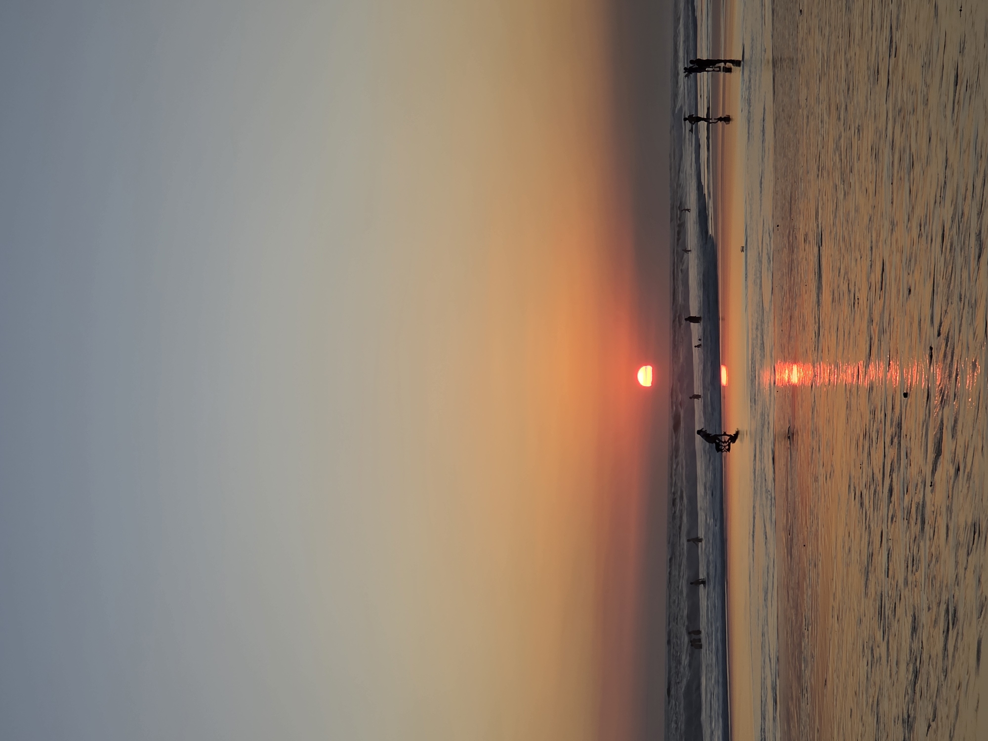The image depicts a sunset at a beach in North Kuta, Indonesia. The sun, a bright orange orb, is low on the horizon, casting a warm glow across the sky and reflecting on the wet sand. Gentle waves are breaking on the shore, with several people visible in the shallow water, wading or swimming. Silhouettes of individuals are scattered along the shoreline, some walking or standing, and one person appears to be engaged in an activity with a dog. The wet sand is smooth and reflective, showing patterns left by the receding tide. The sky transitions from a pale blue at the top to shades of orange and pink near the horizon.