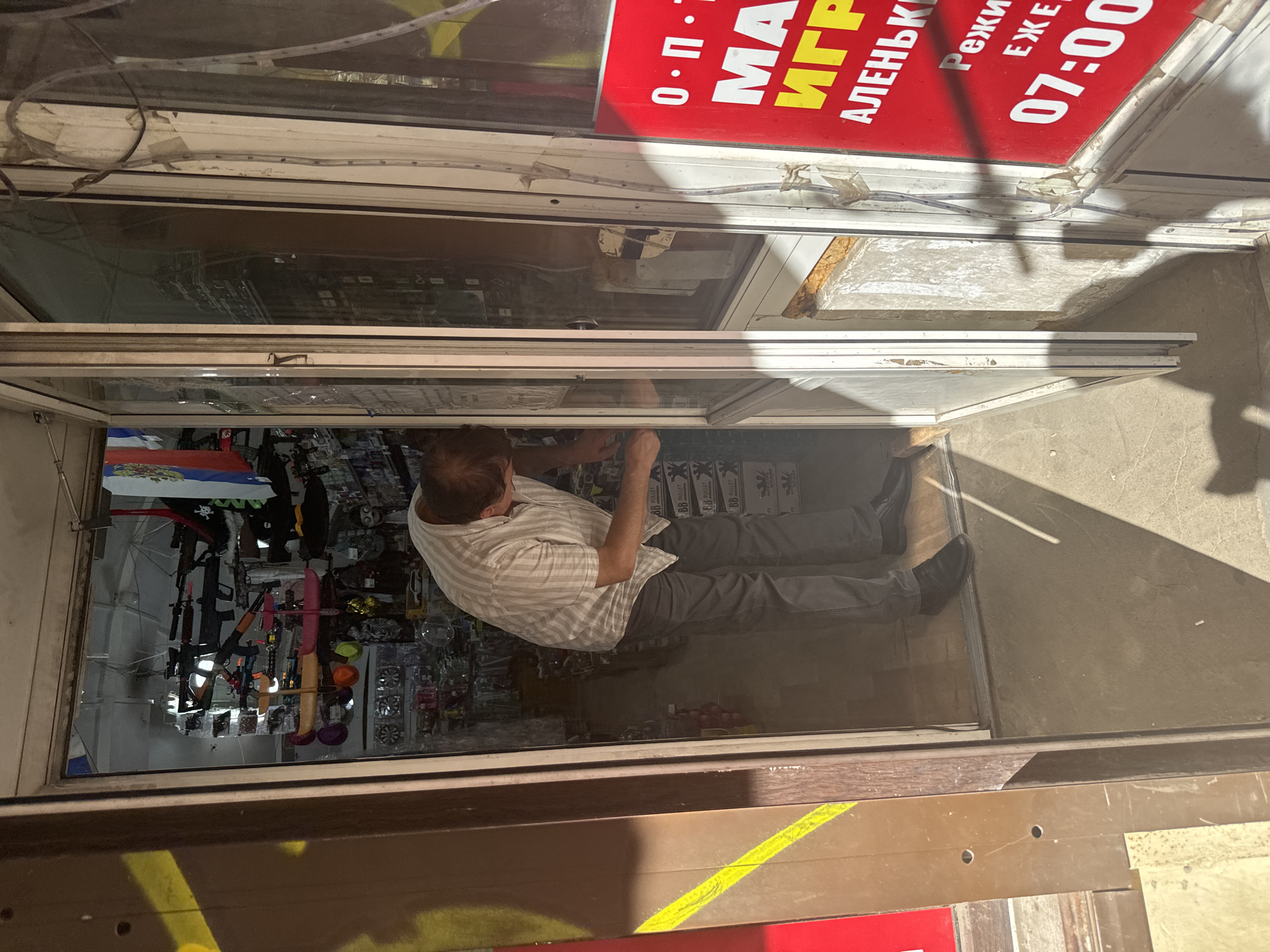 The image shows a man in a light-colored striped shirt and dark pants working at the entrance of a shop. The shop appears to be selling airsoft guns and related equipment, as several are visible hanging on display inside. The shop has a large glass door and a red sign with white lettering that reads "МАИГР" (likely a shortened version of "магазин игрушек" or toy store) and "07:00" indicating opening hours. The shop is located on a street in Saint Petersburg, Russia, as evidenced by the Russian flags visible inside the shop and the inscription on the red sign. The scene is lit by sunlight, casting long shadows. The doorway and surrounding area show signs of wear and tear.