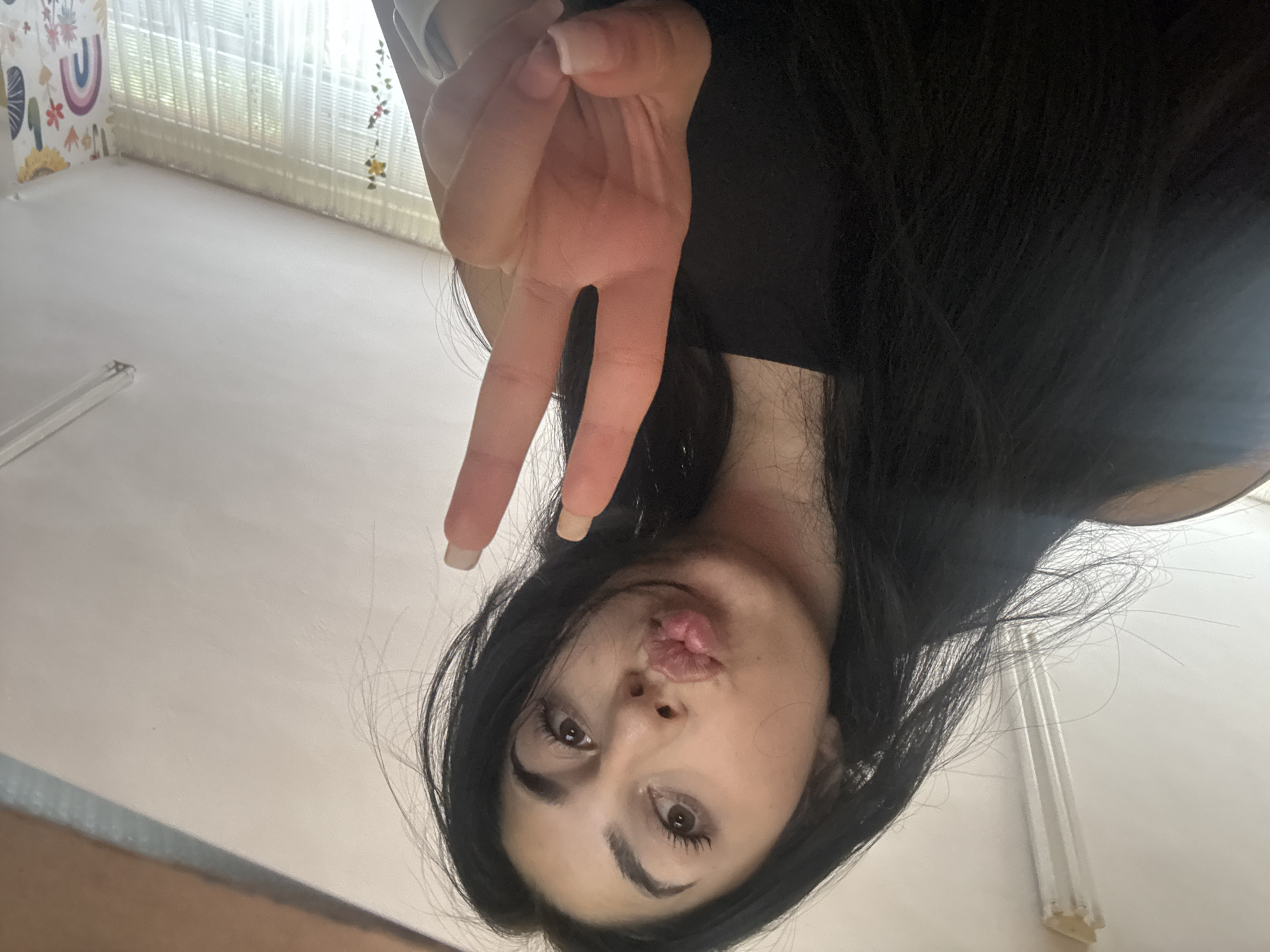 A selfie-style photograph shows a woman with long, dark hair and light skin making a peace sign with her fingers. She is looking directly at the camera with slightly puckered lips. She is wearing a dark-colored top. The background appears to be an interior space, possibly a room with light-colored walls, a window covered with sheer curtains, and decorative elements visible near the ceiling. There is no specific indication of location or landmarks suggesting this is Plovdiv, Bulgaria.