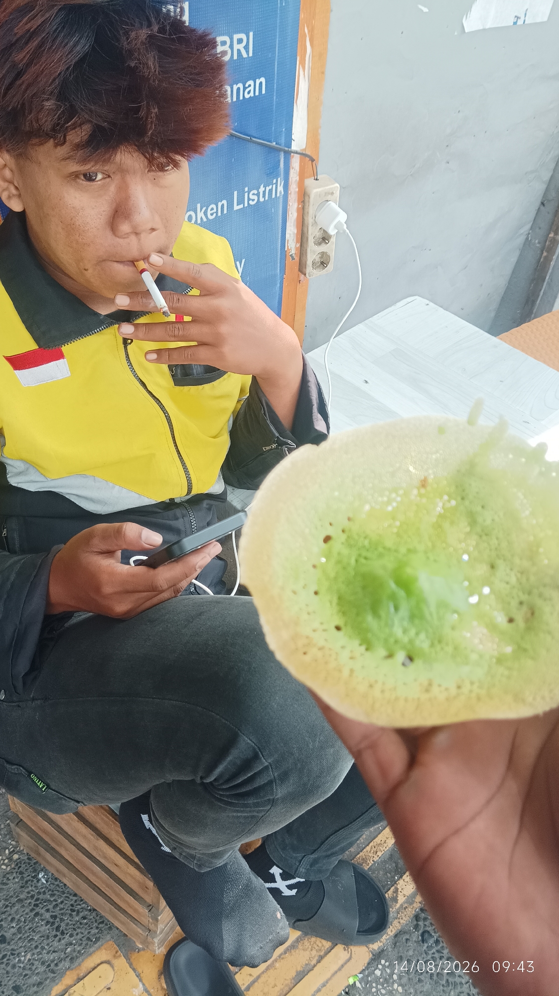 A young man with dark, spiky hair is smoking a cigarette and holding a smartphone in his left hand, while a green pancake-like food item is held in his right hand in the foreground. He is wearing a yellow jacket with a red and white patch on the left arm, dark pants, and black socks with white symbols, paired with black sandals. The background features a blue sign with white text, partially obscured, that reads "BRI" and "Token Listrik." An electrical outlet with a white adapter is visible on the right side of the sign. The setting appears to be outdoors, possibly a street stall, with a rough textured surface and yellow tactile paving on the ground. The image is dated "14/08/2026 09:43".