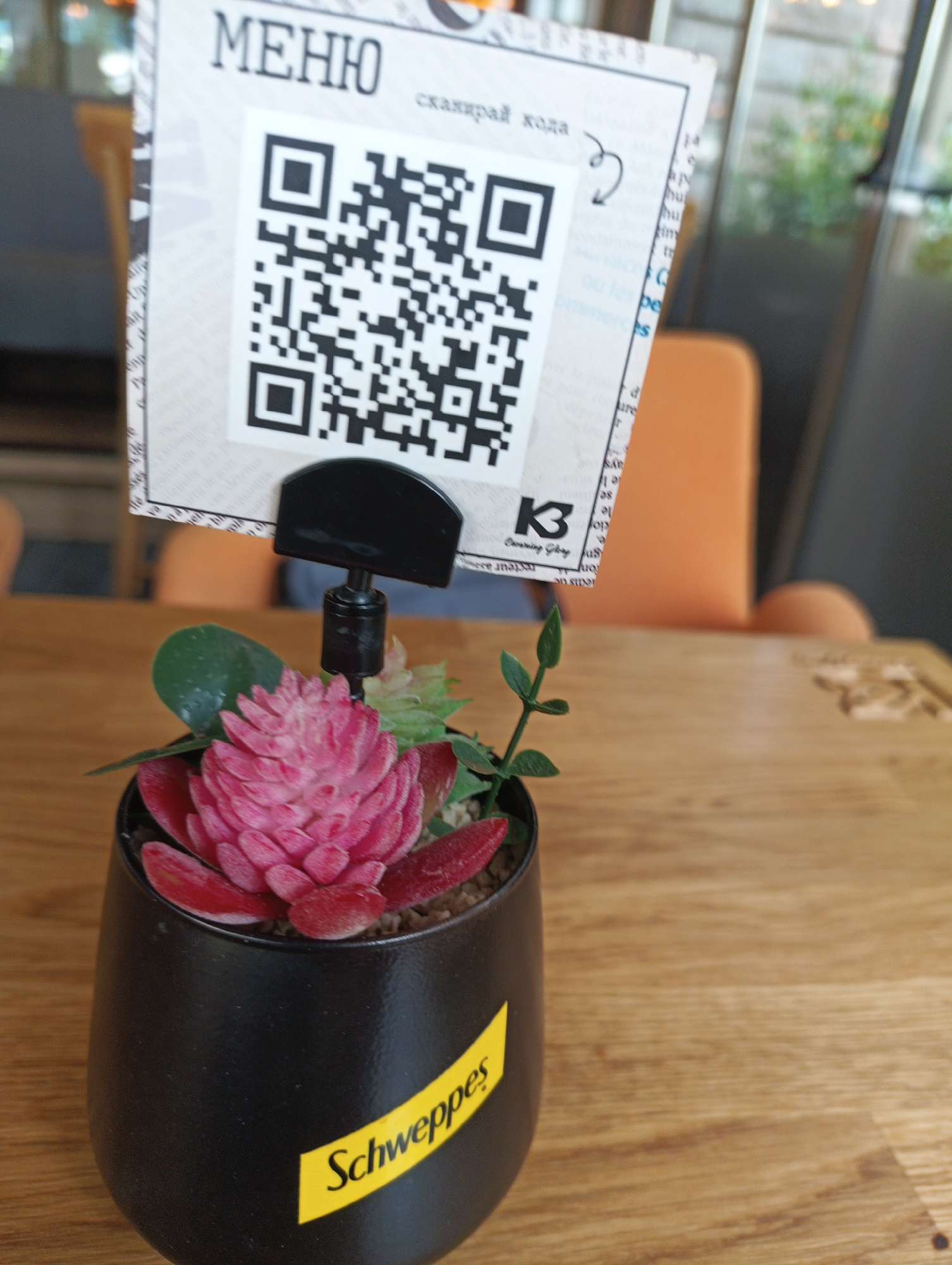 This is a close-up photograph taken indoors, likely in a restaurant or cafe. The central focus is a black pot containing a small artificial succulent arrangement. The pot is branded with a yellow "Schweppes" label. The pot sits on a light brown wooden surface.
Behind the pot is a menu card held by a black stand. The menu card features the word "МЕНЮ" (Menu) at the top, written in Cyrillic script, followed by a QR code. Below the code is the text "сканирай кода," which translates to "scan the code." There is a small logo that is partially visible that looks like the logo for "K3 Coworking Space".
In the blurred background, parts of an orange chair and a glass window are visible. It is possibly located in Burgas, Bulgaria.