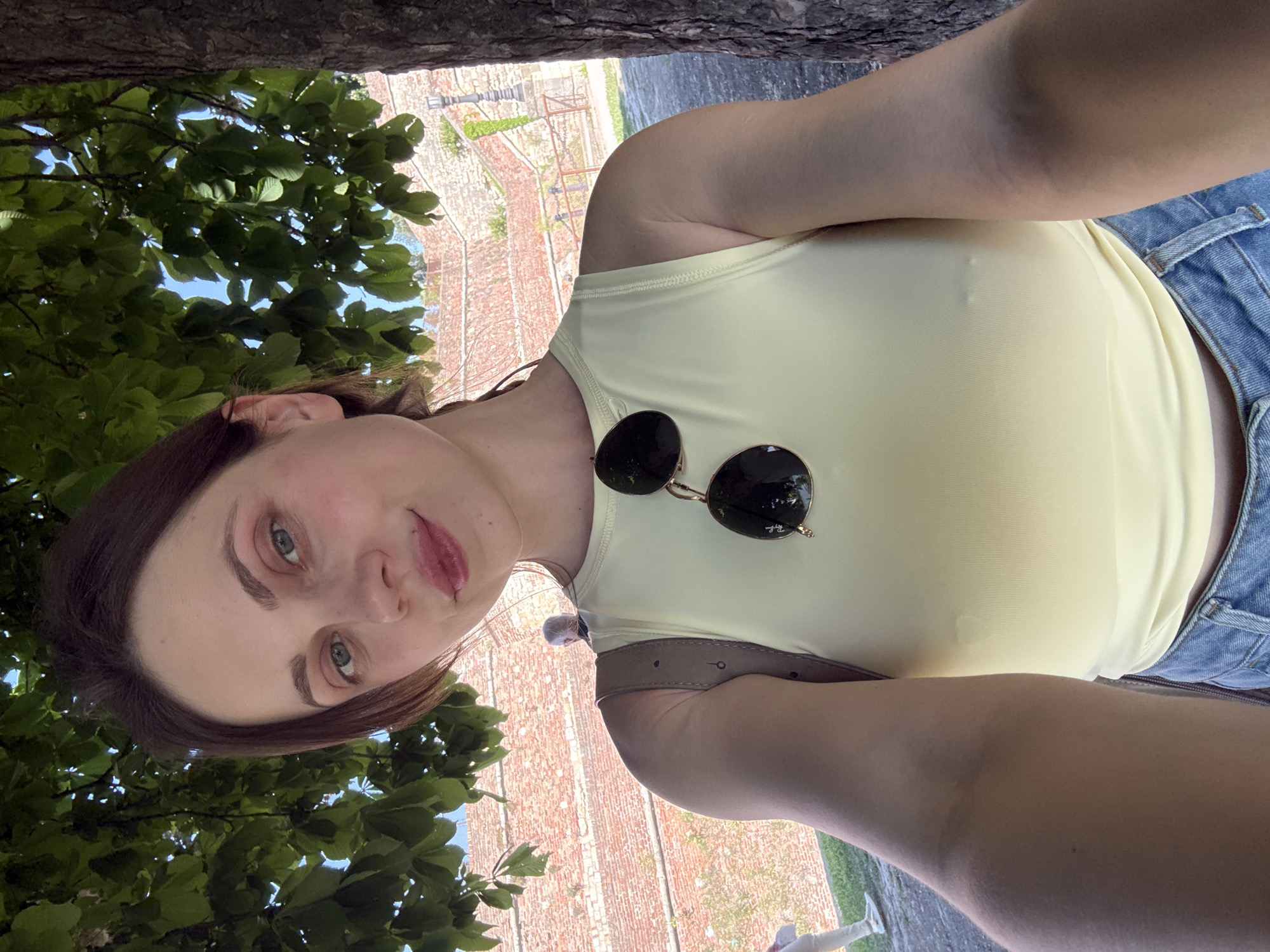 A selfie shot from a low angle features a woman with brown hair and fair skin wearing a yellow top and blue denim shorts. Sunglasses rest on her shirt. In the background, a brick structure is visible, partially obscured by green foliage from a tree. The location is likely a park or garden area in Belgrade, Serbia, given the brickwork's style and the presence of greenery. A tree trunk is visible at the top of the frame.