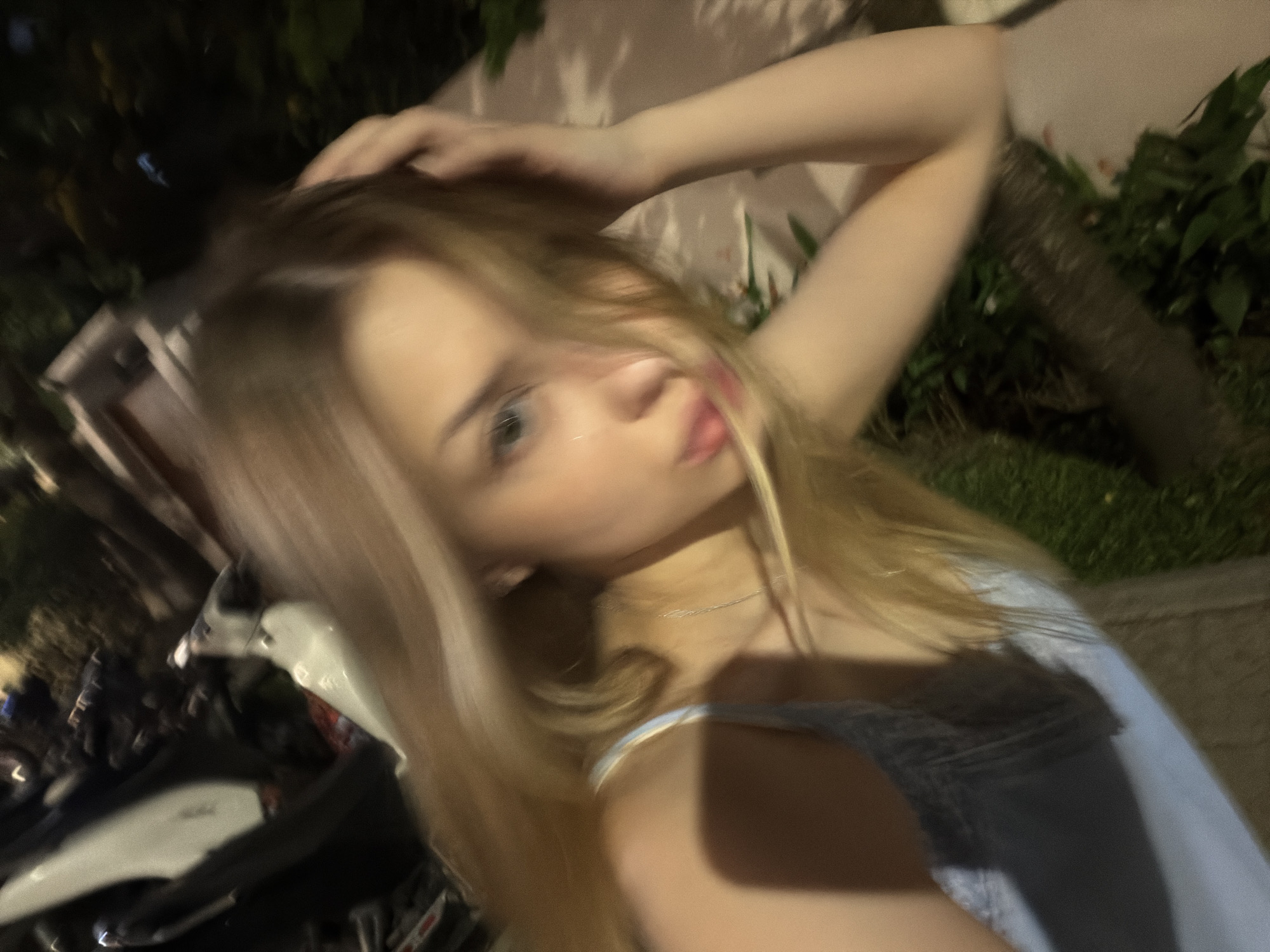 A person with light blondish hair is posing for a blurry selfie-style photograph, likely at night. The person is wearing a light-colored top, and a thin necklace is visible. The left arm is raised, with the hand touching the person's hair. In the background, there's a blurred view of a motorbike parked on the street, some greenery, and a building or wall. It is not possible to precisely identify the location within Đà Nẵng. The details are too indistinct to provide a forensic level description.