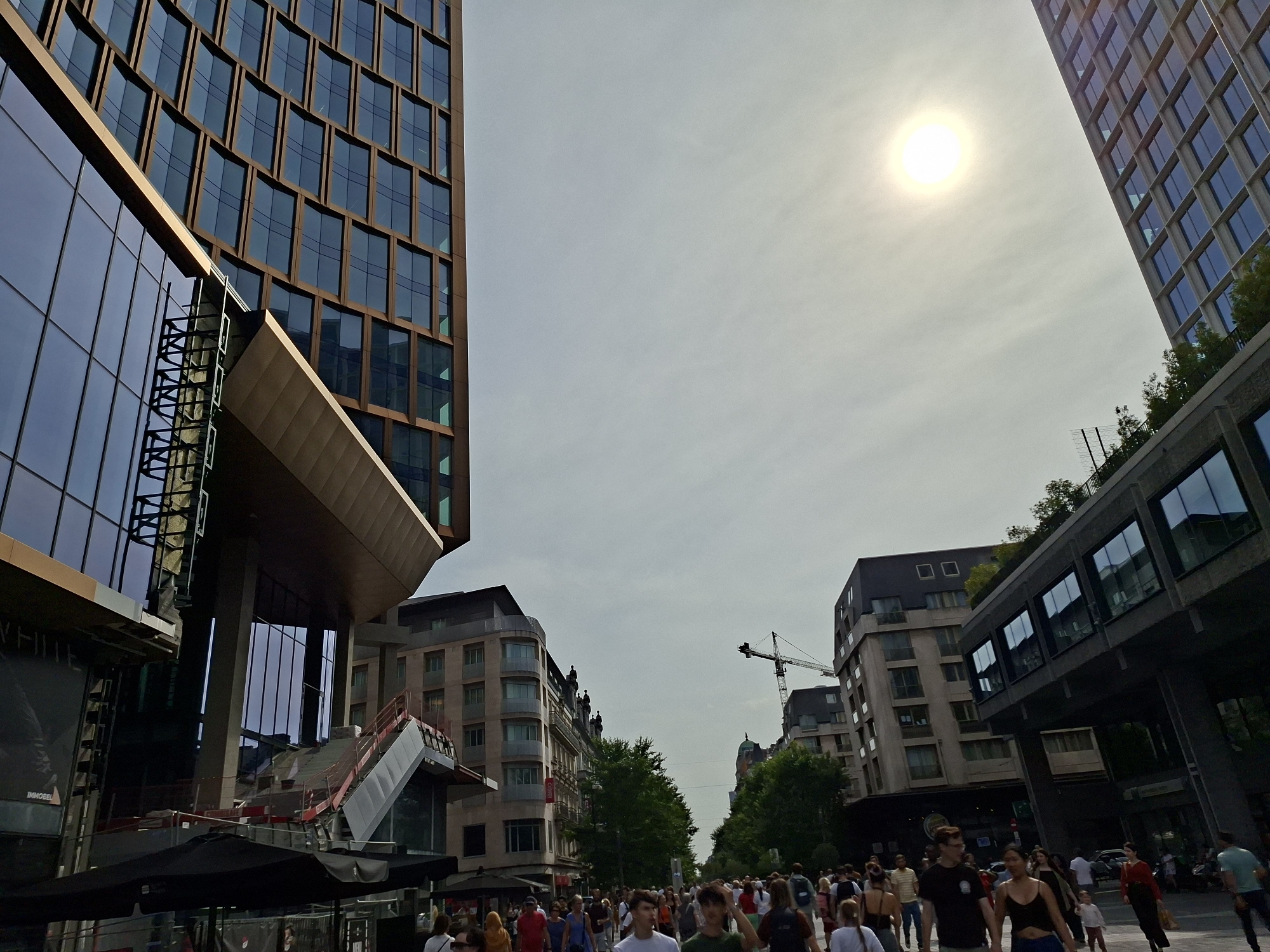 The image captures a bustling urban scene on a bright, overcast day, likely in the city of Brussels, Belgium, given the architectural styles and the presence of a construction crane. In the foreground, a crowd of pedestrians walks along a street lined with modern and older buildings.
Dominating the left side of the frame is a tall, contemporary building with a facade of dark windows set within a bronze-colored grid structure. Below it, a section of the building features a large, reflective glass surface. A large banner is visible on this lower section. Adjacent to this, a partially constructed area shows exposed metal framework and an outdoor staircase with red railings. Several black umbrellas are deployed in this area, suggesting outdoor seating or market stalls.
The right side of the image features another modern building with dark, reflective windows, elevated above street level and supported by a dark, angular structure. Further down the street, older, multi-story buildings with balconies and windows can be seen, interspersed with trees. A construction crane is visible in the middle distance, indicating ongoing development.
The sky is bright with the sun partially obscured by clouds, creating a diffused light. The street is populated by a diverse group of people dressed in casual summer attire, walking in various directions. Their interactions are not clearly discernible due to the distance and focus on the overall scene. The overall impression is of a lively, functional urban environment in the midst of commercial and potentially residential activity.