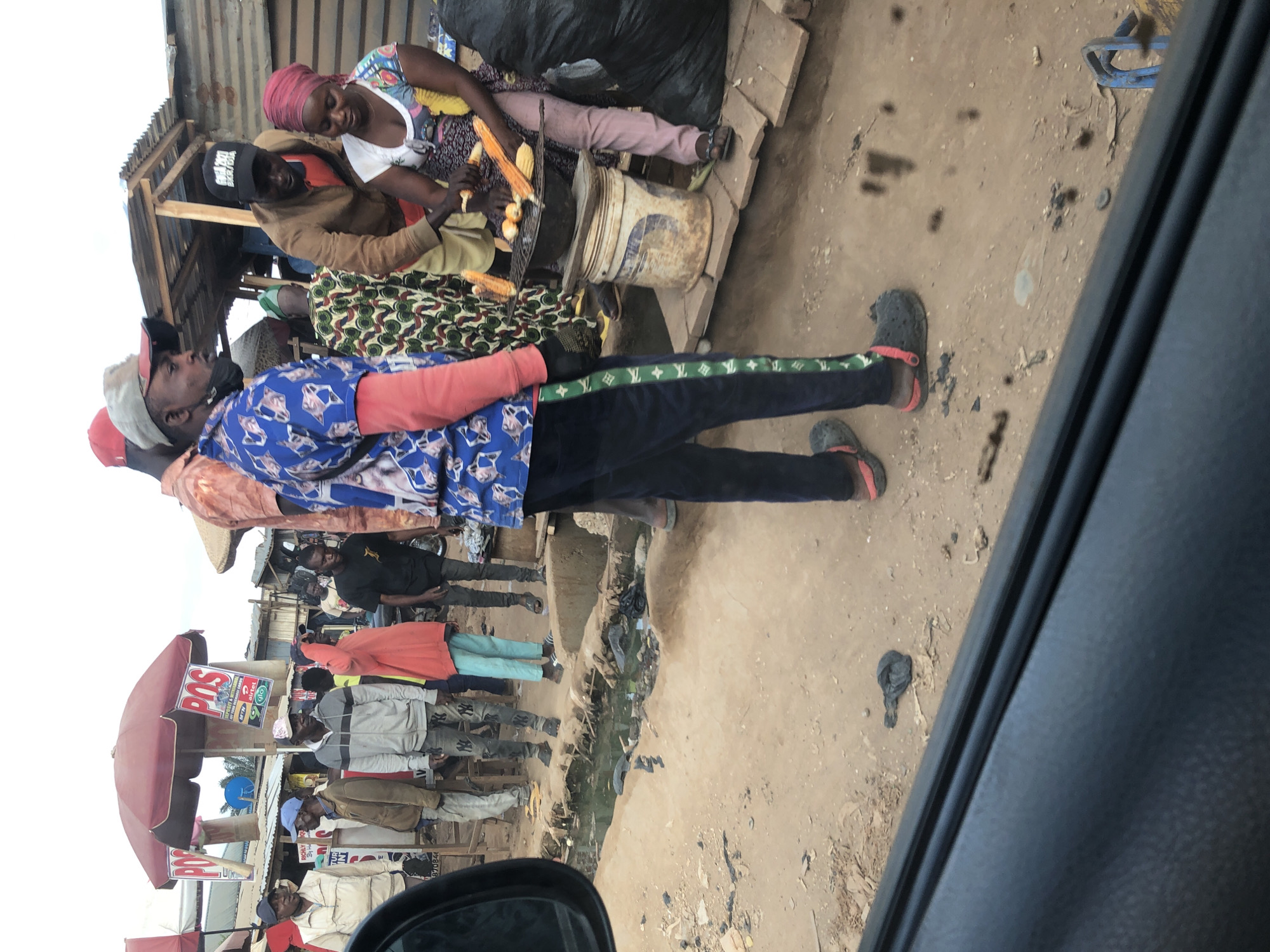 In Otta, Nigeria, a street scene unfolds with several individuals gathered around a food stall. A man in a blue shirt with a repeating pattern is prominently featured, facing left with his mouth open. To his right, a woman in a colorful headscarf and printed dress is preparing corn on a grill. Another man, wearing a brown jacket and a black cap, is interacting with her.
The environment appears to be an outdoor market or street-side area. The ground is predominantly dirt, with some debris and a shallow, stagnant pool of murky water. Several structures, including makeshift stalls with corrugated metal roofs, are visible in the background. One stall has prominent red and white signs advertising "POS" and a telecommunications company. Other individuals, some dressed casually in jeans and jackets, are standing around the area, observing or waiting. The overall impression is one of a bustling, informal urban setting.