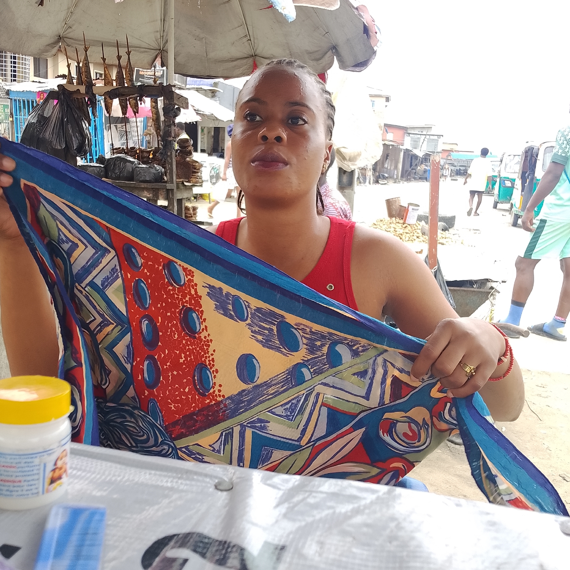 In Ado, Nigeria, a woman is captured in a close-up shot, holding a vibrantly patterned blue, red, and yellow cloth. She wears a red tank top and has braided hair. A small, white jar with a yellow lid is visible in the foreground, to the left of the cloth. The background suggests an outdoor market setting, with various stalls and people. The woman's right hand, adorned with a gold ring and a red beaded bracelet, is partially seen as she manipulates the fabric. The overall scene depicts a moment in a bustling Nigerian marketplace.