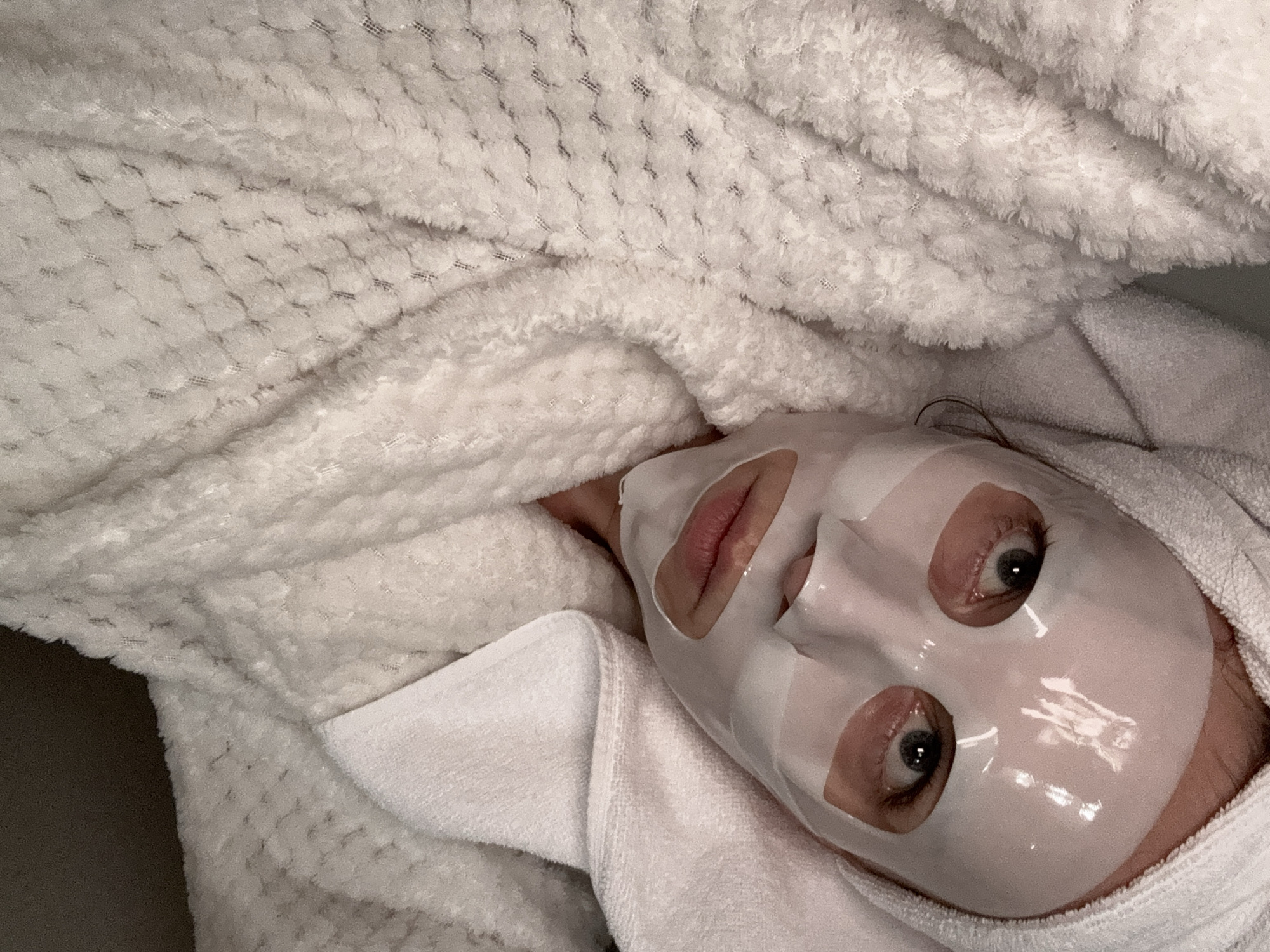This close-up image captures a person engaged in a self-care routine, lying down indoors. The central subject, whose gender is not explicitly discernible, has a white, translucent sheet mask applied to their face. Their eyes are open and looking upwards with a neutral expression, visible through the mask's cutouts, and their lips are slightly parted.
The person is enveloped in soft, white textiles. A plush, white robe or blanket with a textured, possibly waffle-knit pattern, covers most of the frame, creating a cozy and inviting atmosphere. Another piece of white, terry cloth fabric, likely a towel or part of a robe, is visible on the right side and near the subject's neck, further emphasizing comfort. A small strand of dark hair peeks out from beneath the fabric near the ear.
The scene suggests a relaxed moment, possibly in the evening or during leisure time, dedicated to skin care. The lighting is soft and even, contributing to the tranquil mood. While the provided context places this image in Budva, Montenegro, no specific location cues are visible within the frame itself. There is no visible text in the image.