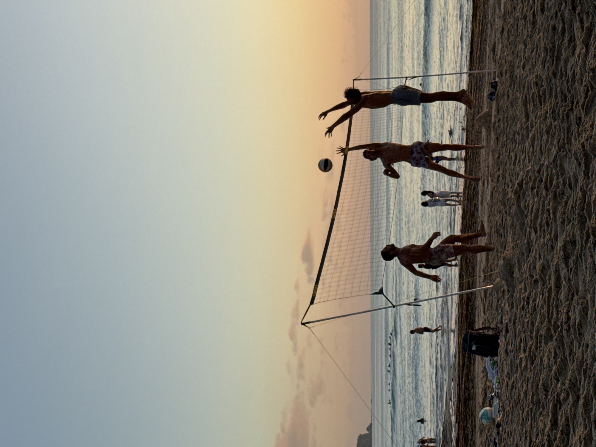 On a sandy beach in Kuta Selatan, Indonesia, during sunset, a group of young men are playing volleyball. The sky displays a gradient of pale blue to soft orange, with the sun low on the horizon casting a warm glow. A volleyball net is set up on the sand, and three men are actively engaged in the game. One man is in mid-air, attempting to spike the ball, while another is jumping to block. A third man is on the left side of the court, having just received the ball. In the background, the ocean stretches out to the horizon, with several people visible wading in the shallow water and a line of surfers further out. Other beachgoers are seen in the distance, enjoying the evening. Scattered on the sand near the volleyball court are a backpack, a volleyball, and some towels. The overall scene depicts a vibrant and active beach environment at dusk.