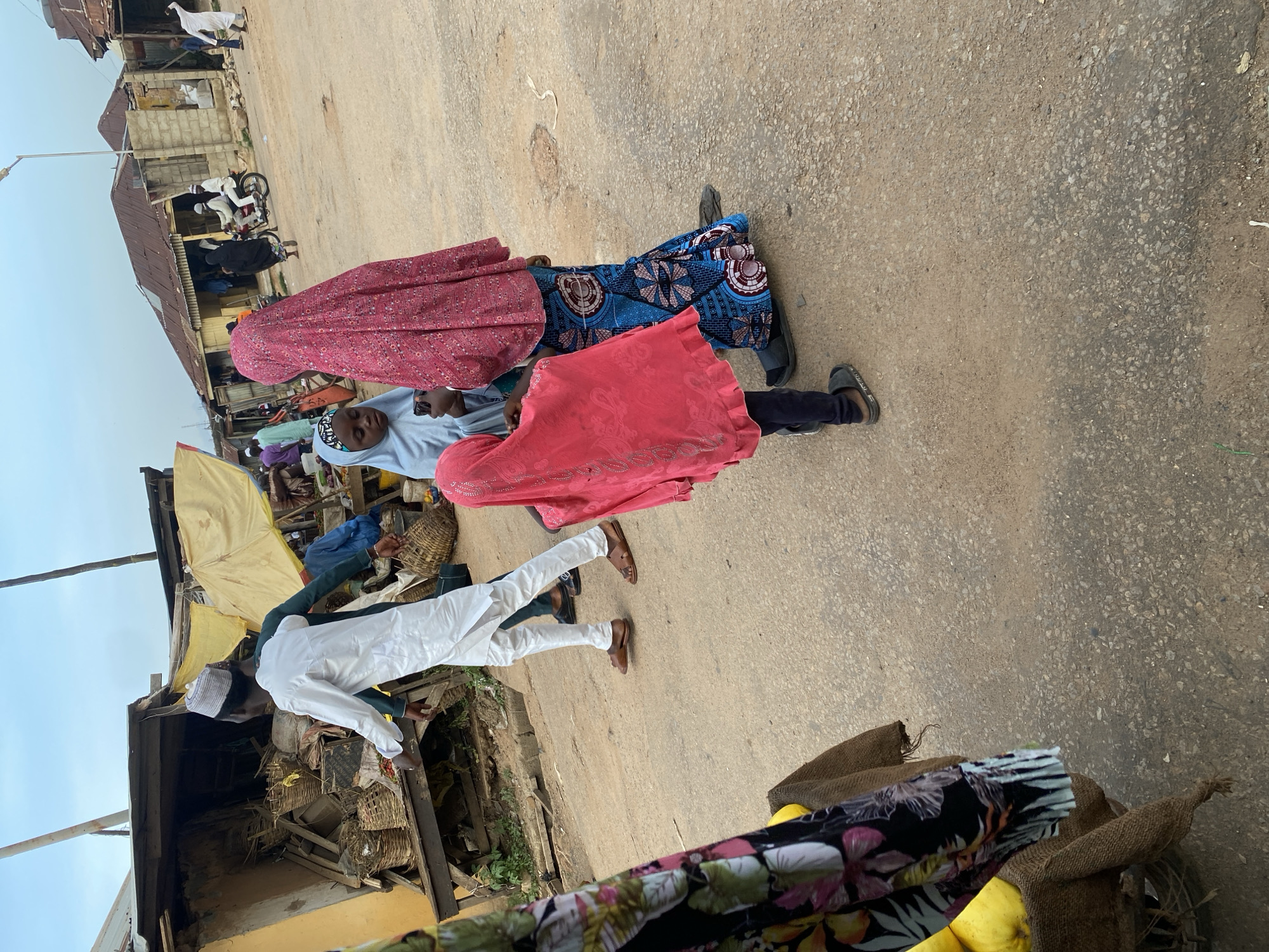 In Jos, Nigeria, a dusty street market is depicted. A man in a white robe and cap walks past a stall overflowing with woven baskets. Two young girls, one in a light blue hijab and the other in a vibrant pink headscarf, stand near an adult woman also wearing a pink headscarf and a patterned blue and white skirt. Behind them, a motorcycle carries two figures. Structures with corrugated metal roofs line the background, suggesting a busy commercial area. The ground is an unpaved, sandy surface.