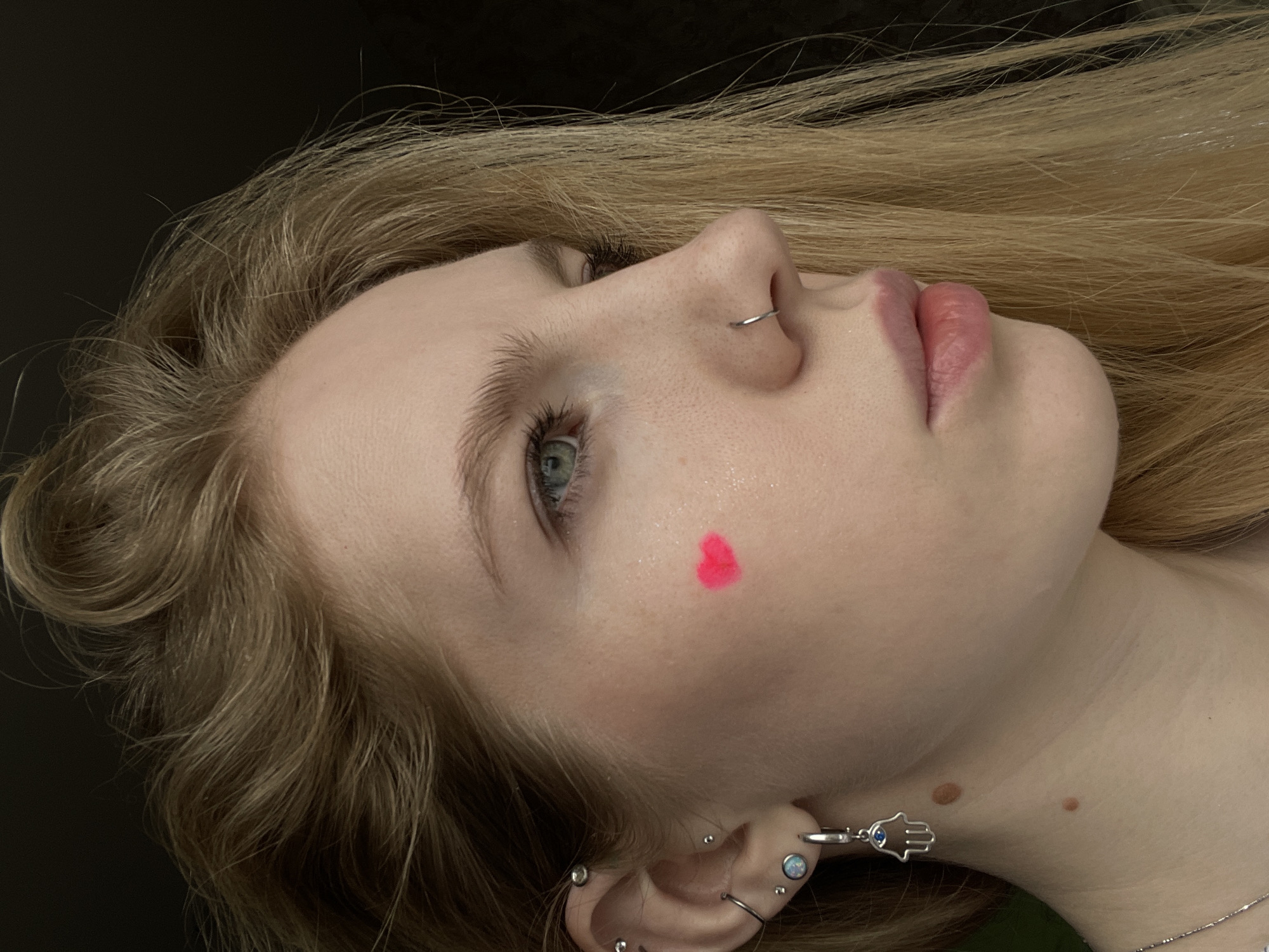A close-up, angled view depicts a fair-skinned young woman with blonde hair. She has a nose ring and multiple ear piercings, including a dangling earring with a Hamsa charm. A bright pink heart is drawn on her cheek. The background is a dark, solid color. There is no indication that this image was taken in Izhevsk, Russia.