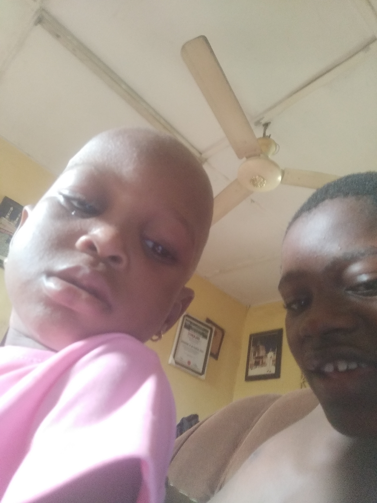 Two individuals, one a child, are positioned closely together, possibly taking a selfie. The child on the left is wearing a pink shirt and has a shaved head. The individual on the right has short hair and is wearing a light-colored garment. A ceiling fan is visible overhead. Certificates or framed pictures are on the wall in the background. The location is in an interior setting, potentially in Shangisha, Nigeria.