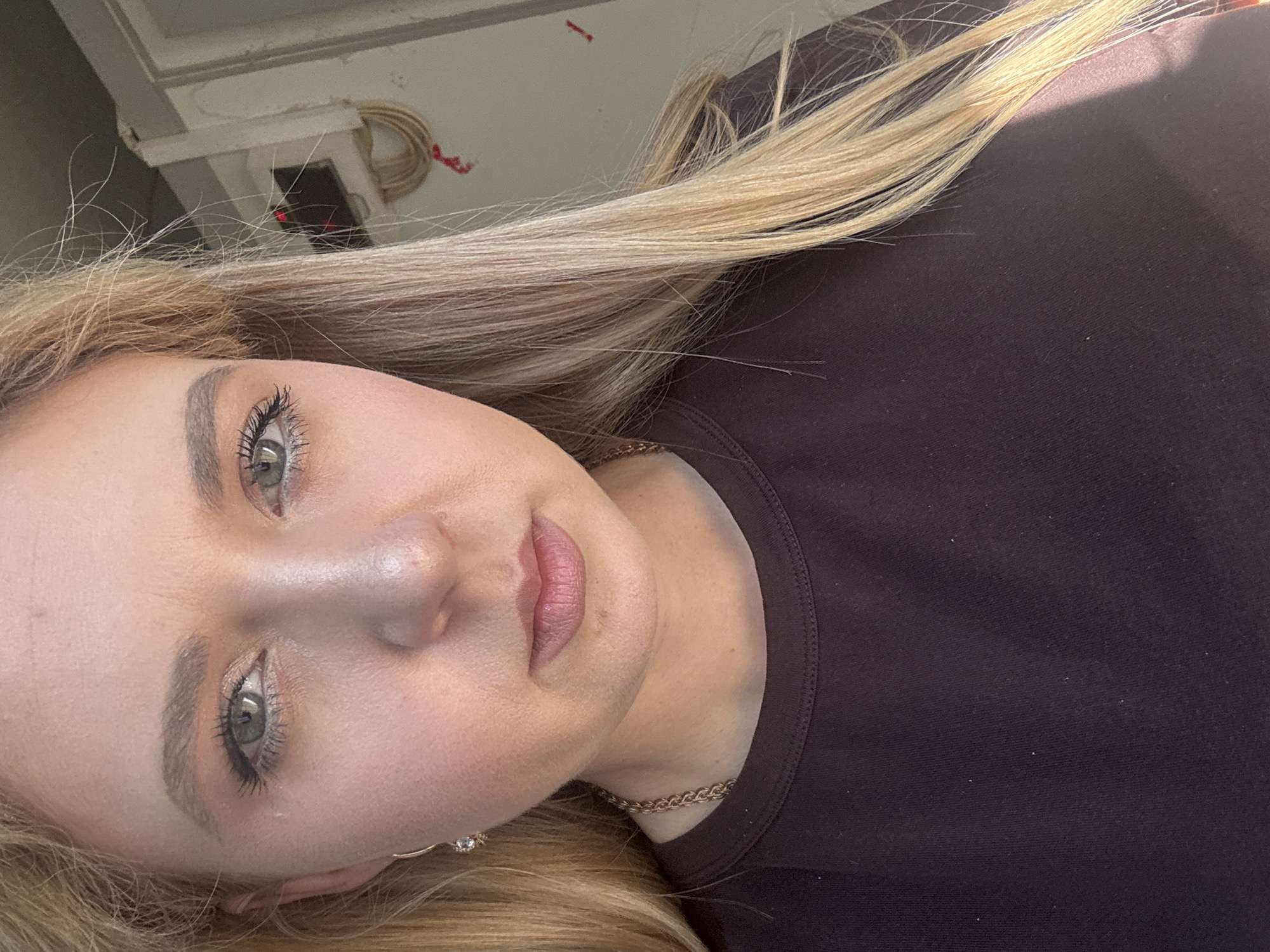 A close-up, waist-up, indoor shot shows a young Caucasian woman from the chest up, facing the camera. She has blonde hair that falls past her shoulders and is wearing a dark purple t-shirt. Her makeup includes mascara, eyeshadow, and lipstick. She is wearing a delicate gold chain necklace and a small gold earring with a stone. The background is blurred, but appears to be a wall with some electrical conduits and possibly a window. The lighting suggests it is daytime.