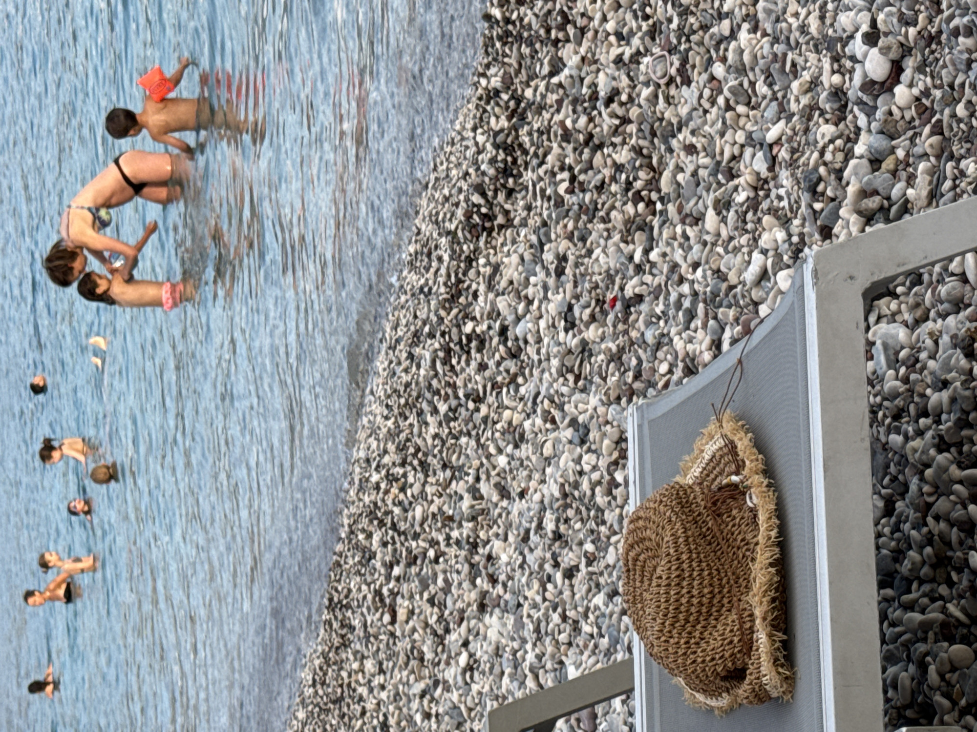 This media file depicts a group of people in the water and on a pebble beach, likely in Budva, Montenegro. Several individuals, including adults and children, are seen wading in the blue water. One person is wearing an orange floatation device on their arm. A straw hat hangs on the side of a grey lounge chair situated on the pebble beach. The scene appears to be a sunny day, suggesting it's daytime, and the presence of people in the water indicates a warm weather activity, such as swimming or cooling off. The water is clear and reflects the sky, while the beach is covered in numerous smooth pebbles of varying sizes and colors. The overall atmosphere suggests a relaxed and leisurely day at the seaside. There is no visible text in the image.