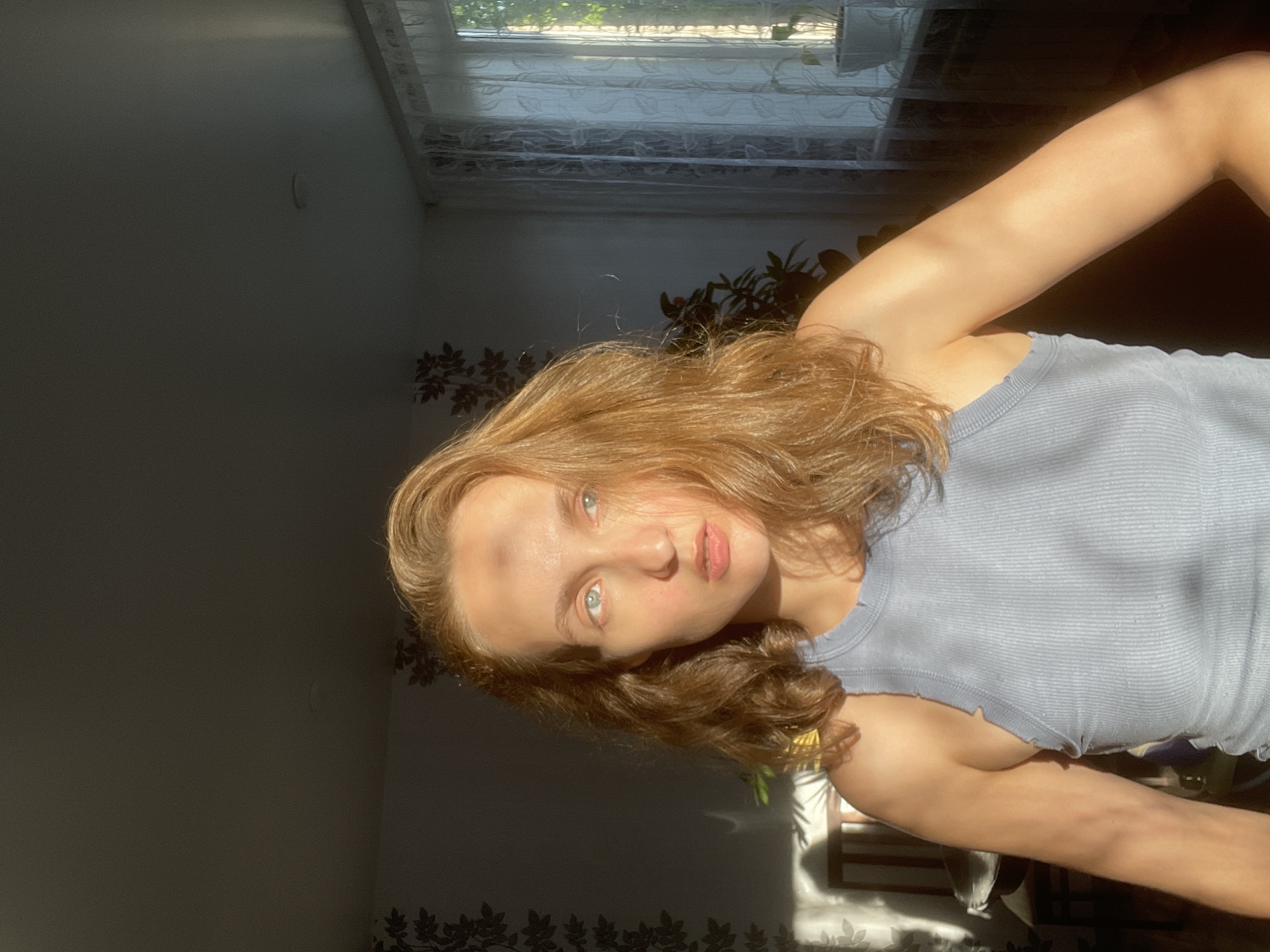 This image, taken in Sharkan, Russia, captures a young woman posing for a photograph indoors. The picture is rotated 90 degrees clockwise, placing the woman horizontally across the frame. She has shoulder-length, wavy, reddish-brown hair and striking light-colored eyes, which appear blue or grey. She is wearing a light blue or grey ribbed tank top with a subtle ruffled hemline. Her expression is calm and neutral as she looks directly towards the camera, suggesting a selfie or a close-up portrait.
The indoor setting is characterized by strong, direct sunlight streaming into the room through a window visible in the background. This intense lighting illuminates parts of her face, hair, and arm, creating a high-contrast scene with bright highlights and deep shadows across the room. This direct sunlight suggests it is daytime, likely morning or late afternoon.
Behind the woman, a window is partially visible with sheer, patterned curtains. Below the window, a houseplant with dark, broad leaves adds a touch of greenery. The wall to the left features a decorative dark floral or leafy pattern along its lower section, while the upper part is a plain, dark grey with a faint, curved mark. The rest of the room remains largely in shadow, emphasizing the subject. No visible text is present in the image.