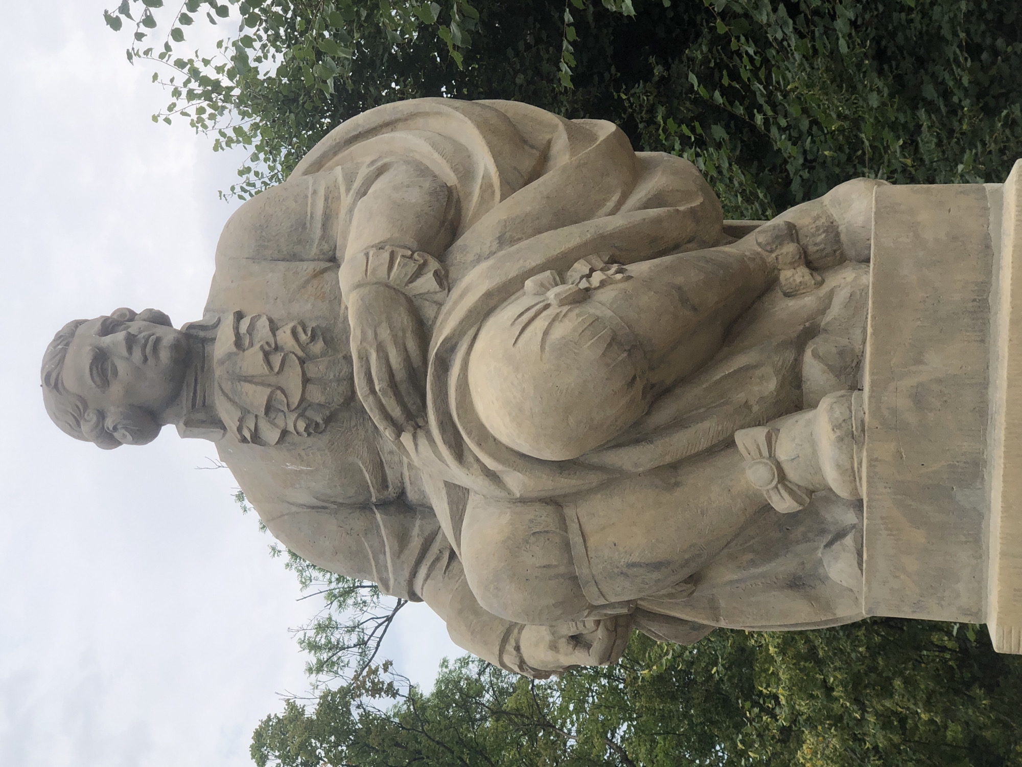 The image shows a sandstone statue of a man in historical clothing. He is seated on a pedestal, with his left leg bent and his right leg extended slightly forward. The man wears a ruffled collar, a long coat, and breeches that are tied with bows at the knees. His hands are clasped in his lap, and he has a pensive expression on his face. The statue is positioned outdoors, with trees and a cloudy sky in the background. The statue is located on the Vistula River escarpment in Warsaw, Poland, overlooking the river.