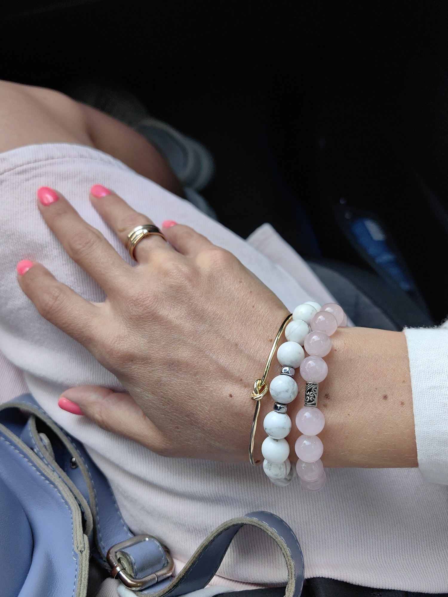 In the city of Янушкавіцкі сельскі Савет, Belarus, a close-up shot focuses on a person's hand resting on a light pink garment, possibly clothing. The hand displays bright pink-painted fingernails and is adorned with a gold ring featuring multiple bands on the ring finger. Two bracelets are visible on the wrist: one composed of white beads with silver accents and a gold knot charm, and another made of pale pink rose quartz beads. A portion of a light blue handbag with gray leather straps and a silver buckle is visible in the lower left of the frame. The background is dark and out of focus, suggesting an indoor or dimly lit environment.