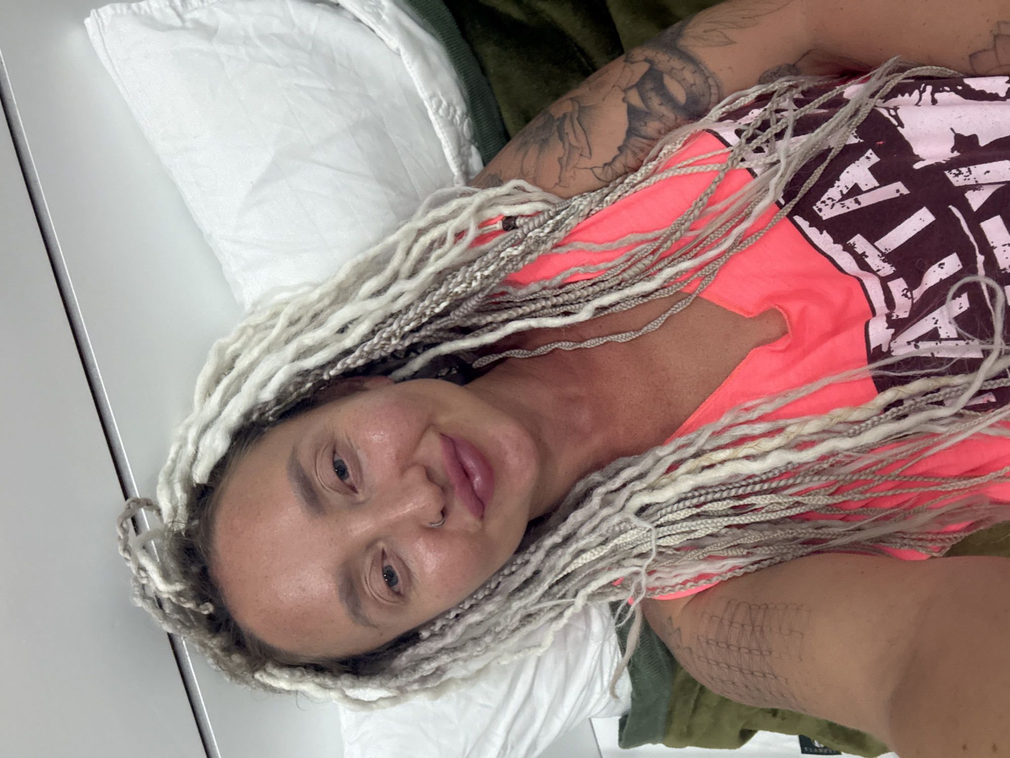 A woman with white dreadlocks and a nose ring lies on a bed with white and green bedding in what appears to be a room in Nha Trang, Vietnam. She is wearing a bright pink tank top with a graphic print. She has tattoos on her left arm and shoulder. The lighting is bright, suggesting daylight or indoor lighting.