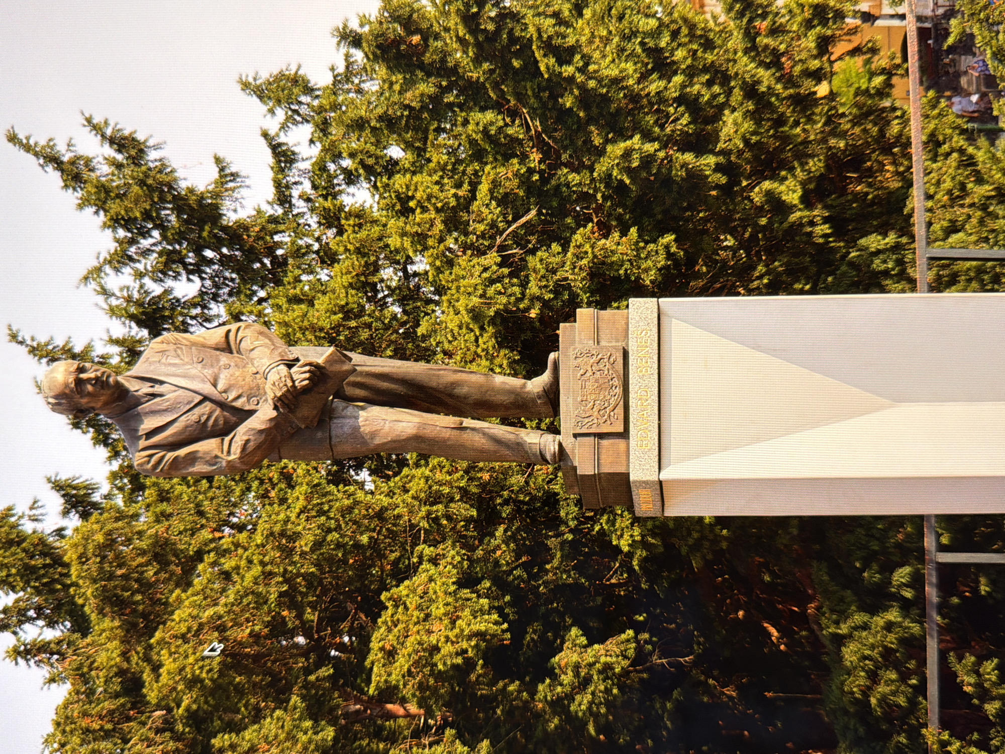 This is a full-body, low-angle shot of a bronze statue of a man in a suit, standing on a tiered pedestal in front of a dense evergreen tree line. The statue depicts a man with short, combed-back hair, a serious expression, and his hands clasped in front of him. He is wearing a suit with a tie. The pedestal is made of stone and features inscriptions and decorative elements. The base of the pedestal is a lighter color and has a geometric design. Behind the statue, the lush green foliage of the trees fills the frame, with some sunlight filtering through. The sky is bright and appears to be a clear day. The statue is located in Kyiv, Ukraine, specifically on Mykhailivska Square. The inscription on the pedestal reads "EDWARD BENES", indicating the statue honors the former President of Czechoslovakia.