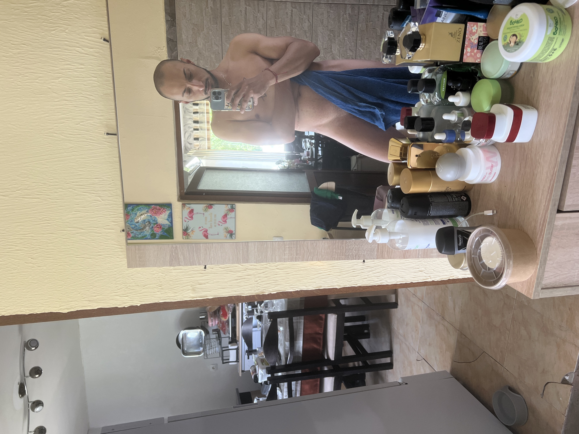 The image is a mirror selfie taken inside a room, possibly a bathroom or bedroom, in Medkovets, Bulgaria. A man is standing in front of the mirror, holding a phone to take the picture. He is partially covered by a blue towel. The room appears to be cluttered with personal belongings. On a shelf in front of the mirror, there are various bottles and containers of toiletries and cosmetics. In the background, a dining table with chairs is visible through an open doorway, suggesting a living or dining area. The overall environment suggests a residential setting.