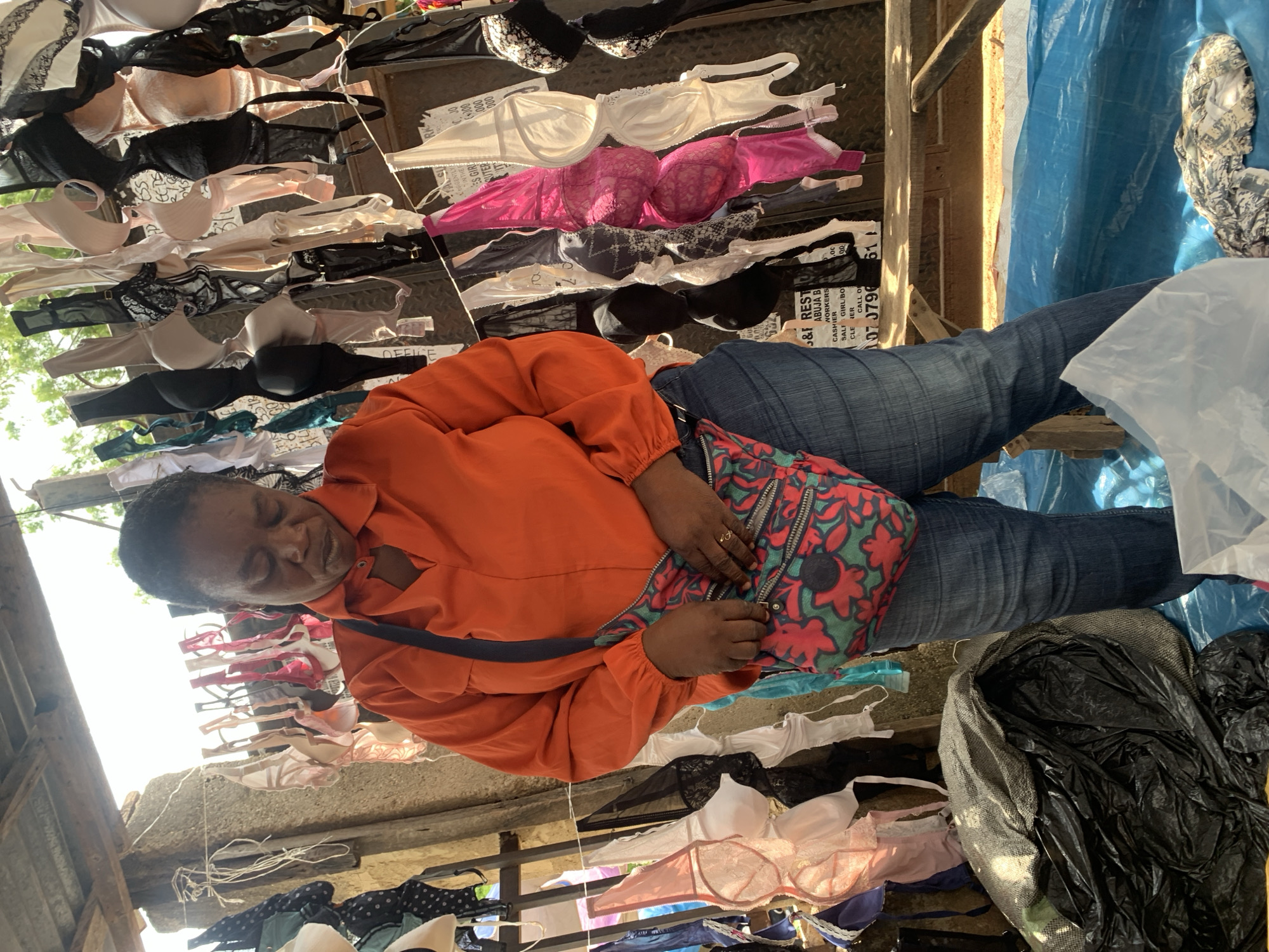 This media file features a woman in an orange jacket and blue jeans, sitting in a market stall filled with bras. The bras are displayed on hangers and lines, showcasing a variety of colors and styles, including white, black, pink, and beige. The setting appears to be an outdoor market in Nyanya, Nigeria, with natural light suggesting it is daytime. The woman has her eyes closed and appears relaxed, perhaps resting amidst the bustling market. A colorful patterned bag rests on her lap, and a ring is visible on her finger. Some plastic bags and other market items are scattered around the scene. Visible text includes "OFFICE," "CASHIER," and what appears to be a phone number. The overall impression is one of everyday commerce in a vibrant market environment.