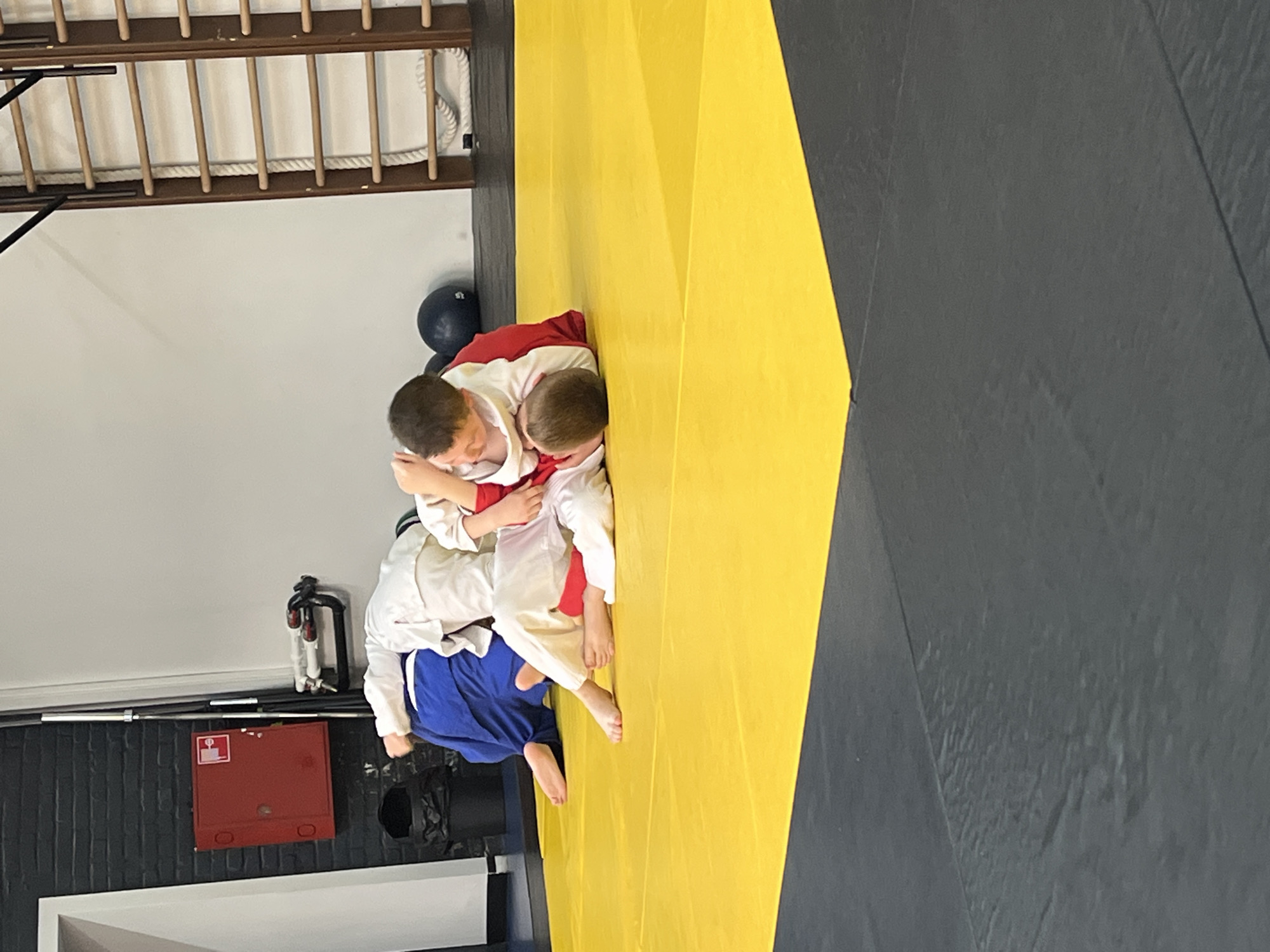 The image shows three children practicing martial arts on a yellow mat in a room with a gray mat and a wall ladder. One child is wearing a white uniform with red trim, another is wearing a white uniform, and the third is wearing a white uniform with blue pants. The children appear to be engaged in grappling. A red fire hose box is visible on the wall, along with a black medicine ball. The location is likely a martial arts studio in Krasnoyarsk, Russia, although this cannot be determined with certainty from the image.