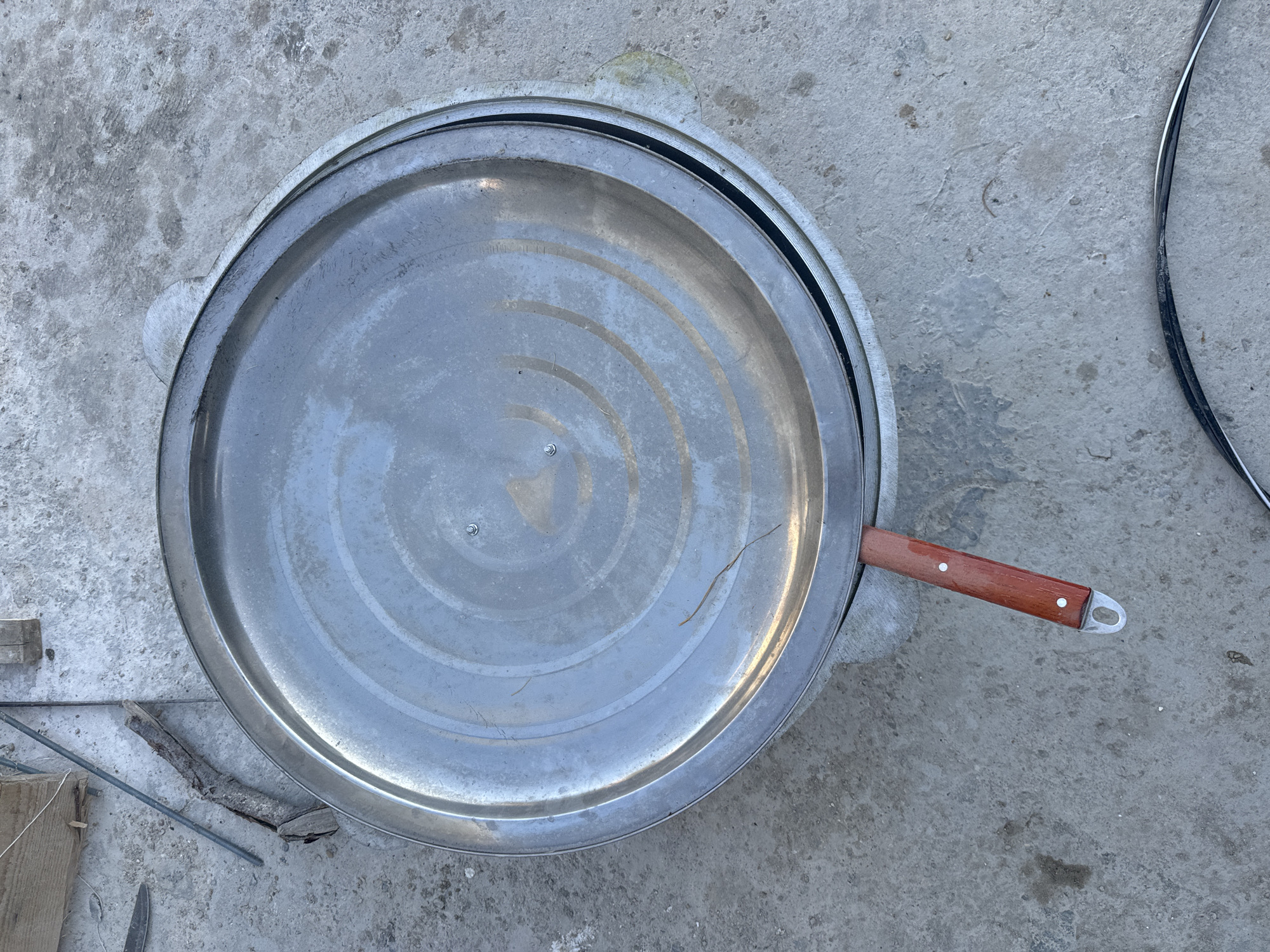 The image shows a large, round, metallic lid resting on a concrete surface. The lid has concentric circular patterns and a small screw visible towards the top right. A wooden handle with two rivets is attached to the rim of the lid and extends outwards. In the background, a piece of wood and some metal rods are visible. The scene appears to be outdoors on a rough concrete ground. The location is identified as Atameken, Kazakhstan. There are no people or interactions depicted.