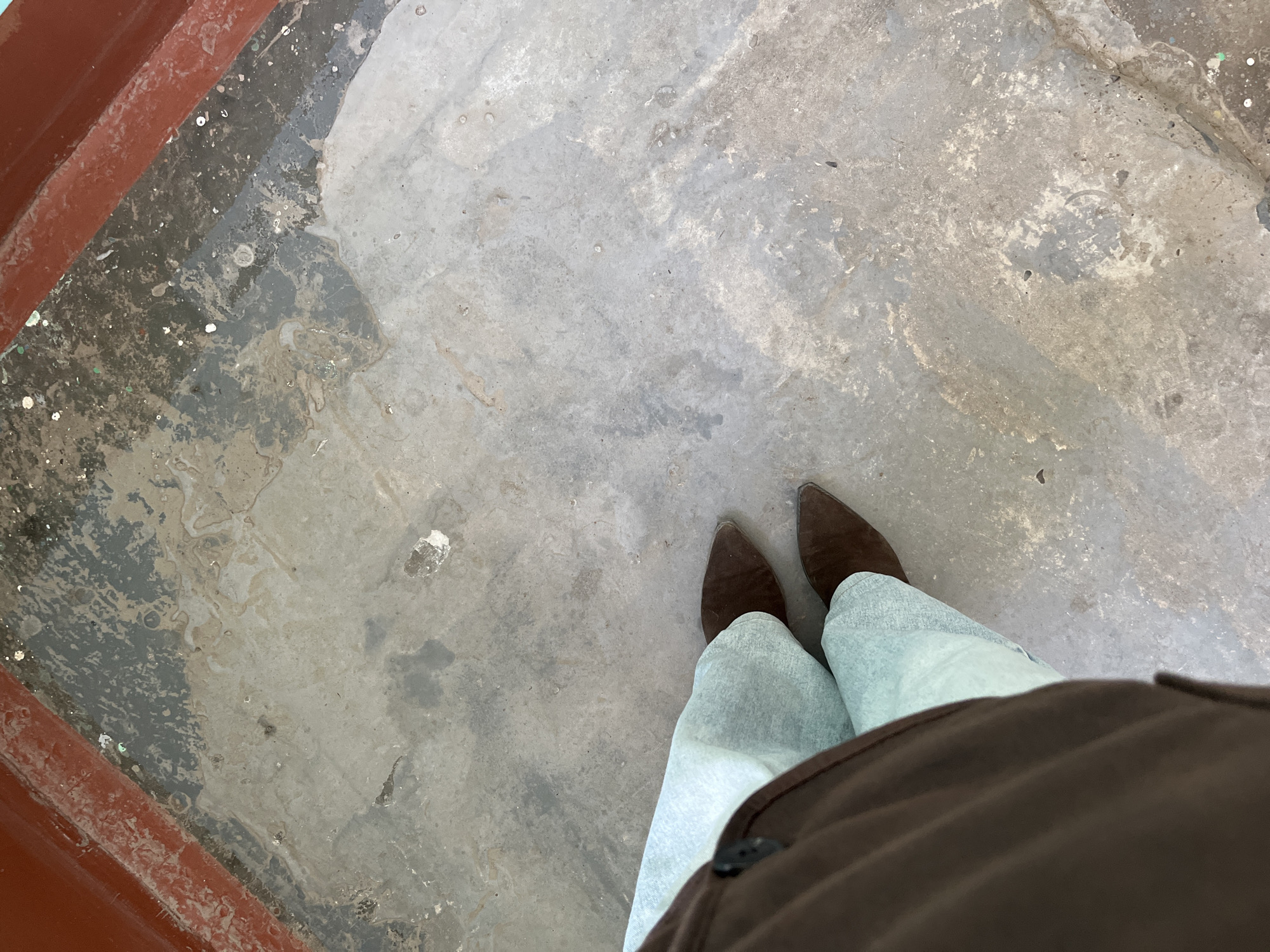 A high-angle shot depicts a person standing on a worn, gray concrete surface. The person is wearing light-colored denim jeans and brown, suede-like pointed-toe boots. A portion of a brown garment, possibly a jacket or coat, is visible in the lower right corner of the frame. The concrete surface is uneven and appears weathered, with patches of darker gray and some debris. In the upper left corner, a section of red-brown paint or metal is visible. The location is not evident from the visual details provided. It is not possible to determine that the image was taken in Chaikovsky, Russia.