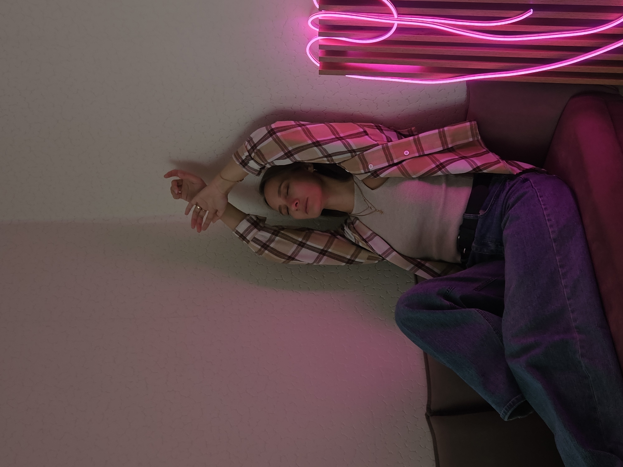 An indoor scene depicts a light-skinned female seated on a dark brown couch with a dark red cushion. She leans against a textured, light-colored wall with her eyes closed and head tilted back slightly. Her arms are raised and crossed above her head, resting against the wall. She wears a light-colored ribbed tank top beneath an open long-sleeved plaid shirt with tan, brown, white, and pinkish-red checks, and blue denim jeans. A thin gold necklace and another with a small cross pendant are visible around her neck. A gold ring is on her right ring finger. To her right, a bright magenta neon light, shaped as abstract curves, is mounted on a vertical wooden slat panel. The scene is illuminated by ambient light and a prominent pink/magenta glow originating from the neon fixture, reflecting on the left wall and the subject. The walls exhibit a textured pattern resembling cracked plaster or stucco. The specific geographic location in the city of Chaikovsky, Russia, cannot be determined from the visual information provided within the image.