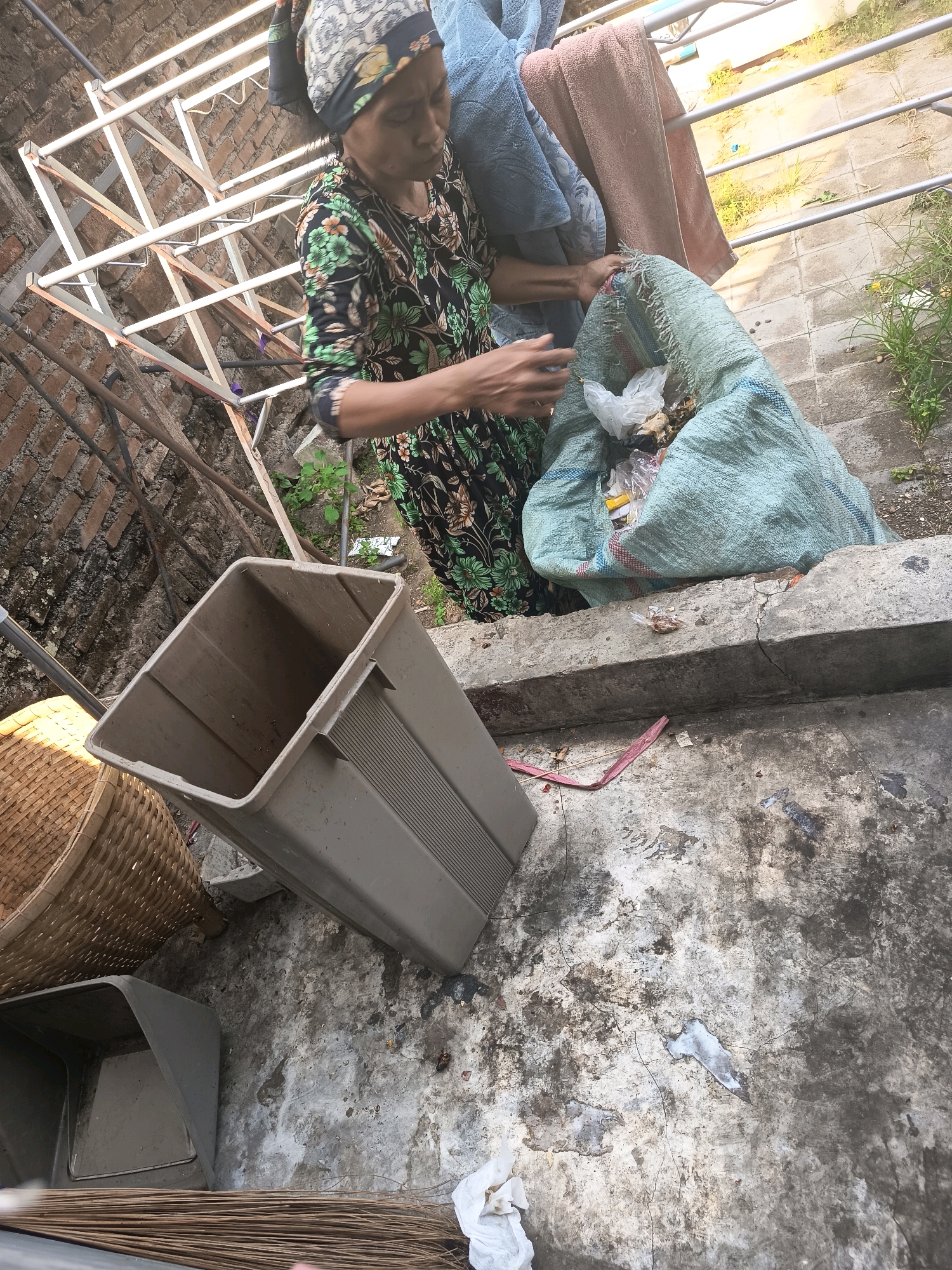 In Banjar, Indonesia, an elderly woman with a headscarf, wearing a floral dress, stands near a clothesline holding a large, light blue woven bag filled with trash. Beside her, to the left, is a large, beige plastic bin, and to the far left, a woven basket. The scene takes place outdoors on a concrete surface, with a brick wall visible in the background, suggesting a residential area. The ground is littered with debris.