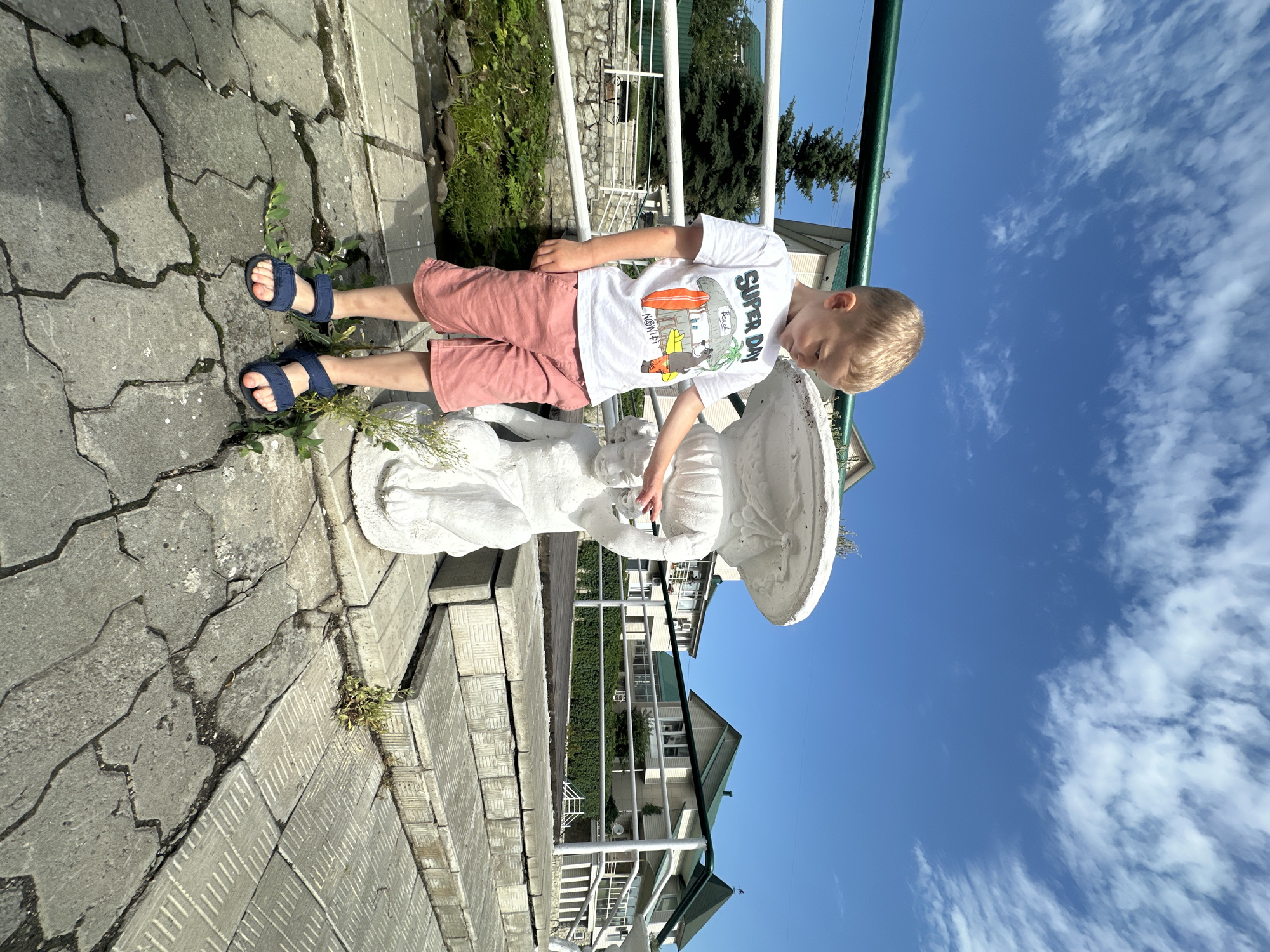 In Даниловка (алтайский), Russia, a young boy with blond hair stands on a paved walkway next to a white, child-like statue that appears to be part of a fountain. He is wearing a white t-shirt with a graphic print and coral-colored shorts, along with dark blue sandals. The boy is touching the statue with his right index finger. The background shows green trees, white buildings with green roofs, and a clear blue sky with scattered clouds. A green railing runs along the edge of the walkway. The ground is made of grey interlocking pavers, some of which are cracked and overgrown with small plants. The lighting suggests it is daytime, likely sunny.