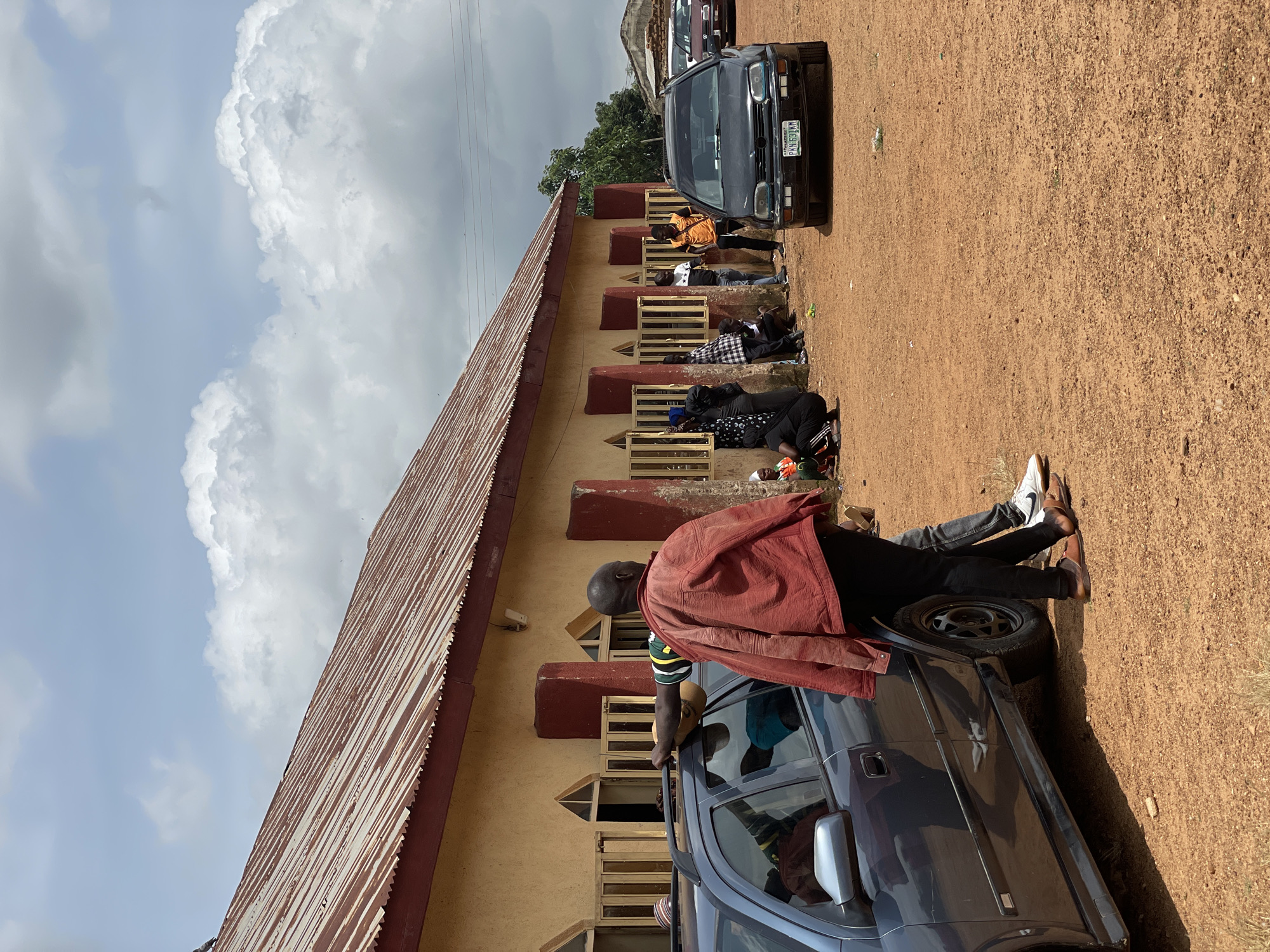 An outdoor, eye-level, medium shot captures a group of people standing and sitting outside a long, single-story building with a corrugated metal roof. The building's exterior walls are painted a pale yellow with dark red columns at intervals. Windows with barred openings are visible along the building's facade. The ground is covered in reddish-brown dirt.
In the foreground, a dark grey car is parked on the left side of the frame, facing right. A man in a red jacket and black pants stands to the right of the car, leaning on its roof. His back is to the camera.
To the right of the car, a black sedan is parked facing the camera. Its license plate reads "PKN 631 KM". Other cars are visible further in the background.
A group of people are gathered near the building, some standing, some sitting on the ground. They appear to be engaged in conversation or waiting. The sky above is partly cloudy. The location is Kassa, Nigeria.
