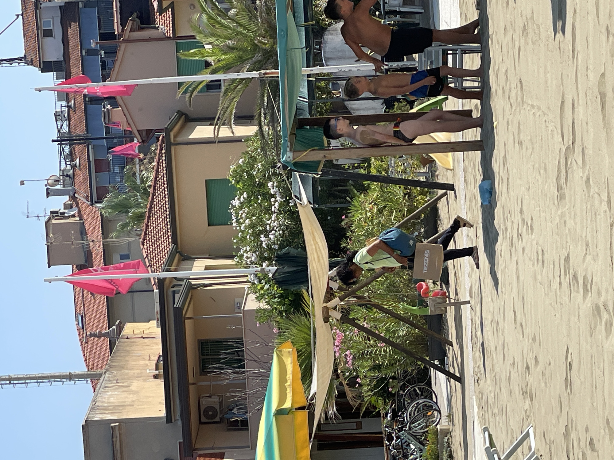 The image depicts a sunny day at a beach in Viareggio, Italy. In the foreground, sand stretches across the scene, with a few white beach chairs partially visible on the left. A group of people, including several young boys and one woman, are present. The woman, wearing a lime green t-shirt and black pants, is walking and carrying a shopping bag that reads "TEZENIS." She has a blue bag slung over her shoulder. The boys are dressed in swimwear and appear to be standing near a shaded area with a green awning and wooden posts. Behind them, lush green foliage and palm trees are visible, along with buildings with terracotta roofs and some flags, one of which is pink, flying in the background. A yellow and green umbrella is also present on the left side of the image. Bicycles are parked on the left, suggesting the presence of a bike path or rental area. The overall atmosphere is one of a casual beach day.