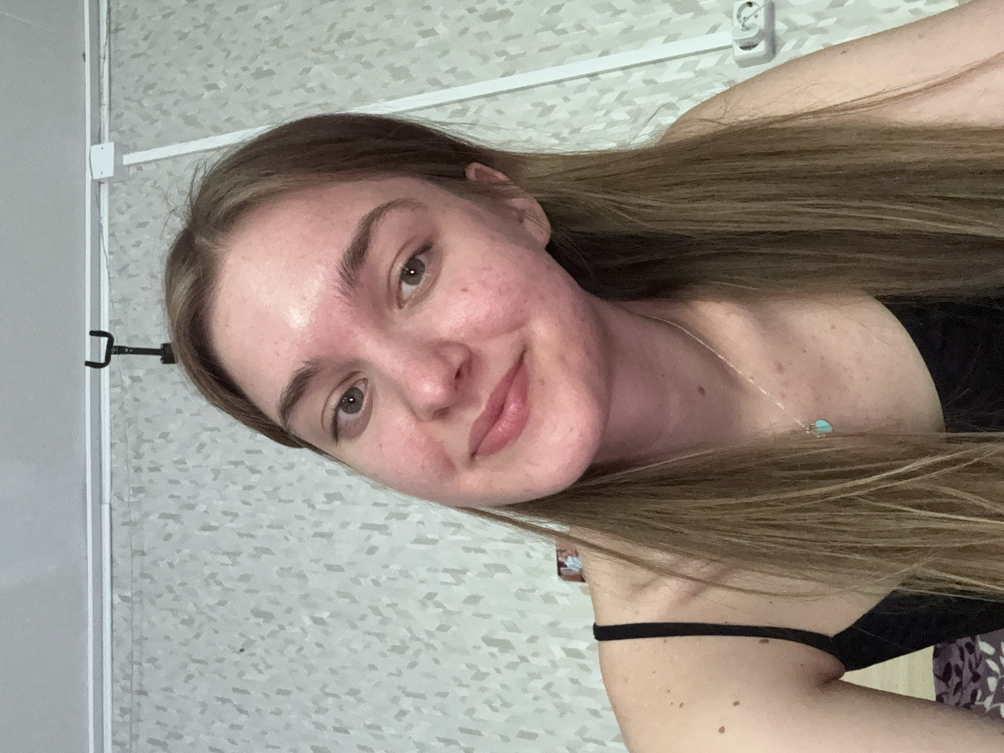 A young woman is pictured in a selfie, taken indoors. She has long, light brown hair, fair skin, and is wearing a black top. A silver necklace with a light blue pendant is visible. The background features a light-colored wall with a patterned design, and a white cable or pipe running horizontally along the top. A white electrical outlet is visible near the top right. There is also a black holder of a mobile phone. The location in Perm, Russia, is not evident in the image.