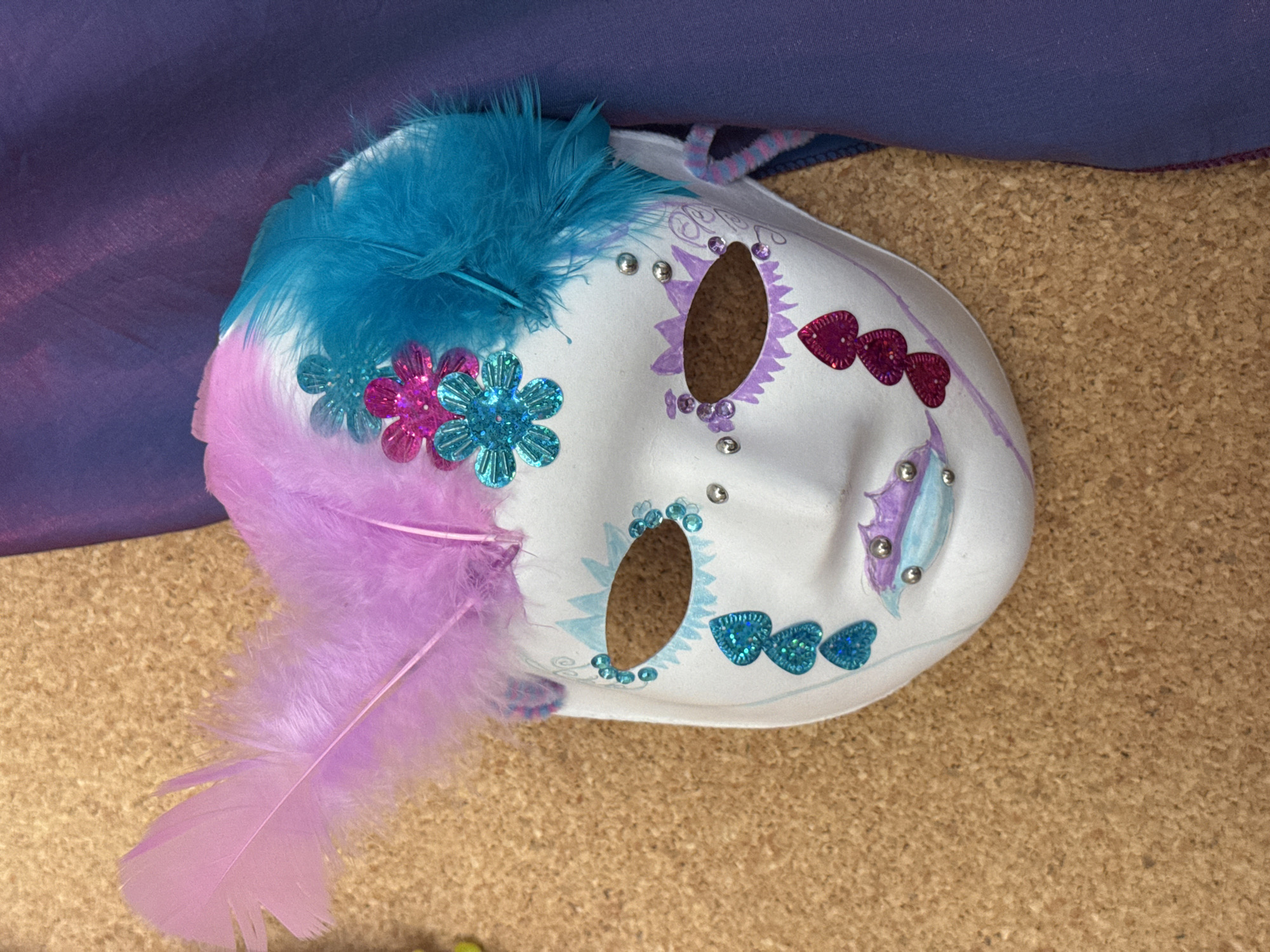 A decorative, white mask is the central subject of the image, positioned on its side, angled towards the bottom right. The mask is extensively adorned with colorful embellishments.
On the left side, a cluster of soft, light purple feathers extends downwards, while a prominent tuft of teal/turquoise feathers sits at the top left edge of the mask. Below the teal feathers and to the left of the eye opening, there are two glittery, sequined flower appliques: one in magenta and one in teal. Another pair of magenta and teal sequined flowers are positioned slightly further down.
The mask features oval-shaped eye openings, revealing the textured cork board surface beneath. Around the right eye opening (from the mask's perspective), there are drawn purple swirled designs, accented with small purple rhinestones and silver studs. Above this eye, three magenta glittery heart appliques are arranged in a row. Similarly, around the left eye opening, light blue drawn designs are visible, accompanied by small teal rhinestones and silver studs. Below this eye, three teal glittery heart appliques are arranged. A subtle light purple outline defines the edges of the mask. A braided strap, featuring pink, blue, and purple colors, is partially visible near the top right edge of the mask.
The background is divided into two parts: a dark purple fabric covering the upper left portion of the image, and a textured cork board surface occupying the bottom right. The mask appears to be stationary, possibly displayed or awaiting use. No activity is taking place, and there is no visible text within the image.